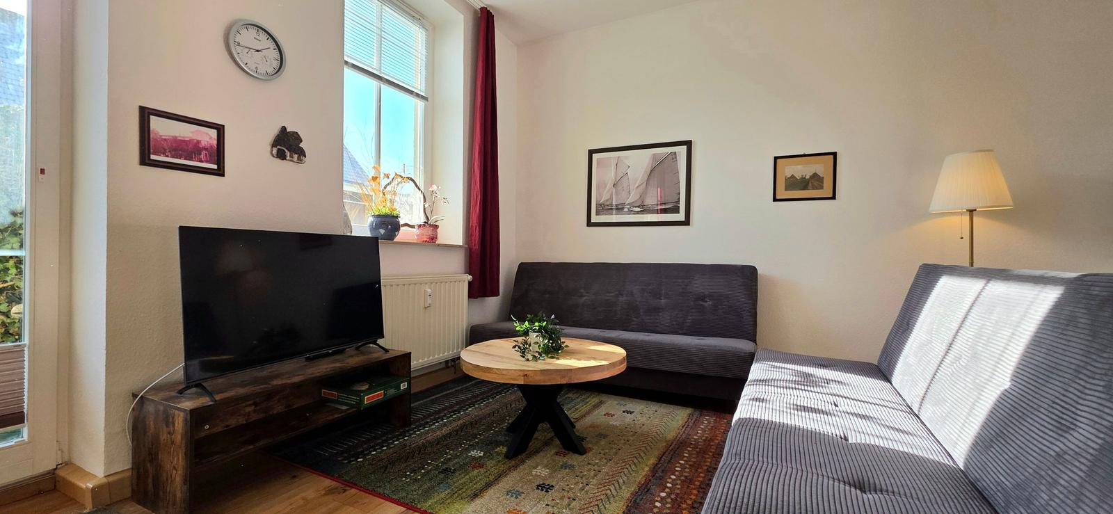 Living room with sofa, TV, and table. Window with curtains. Wall pictures and lamp.