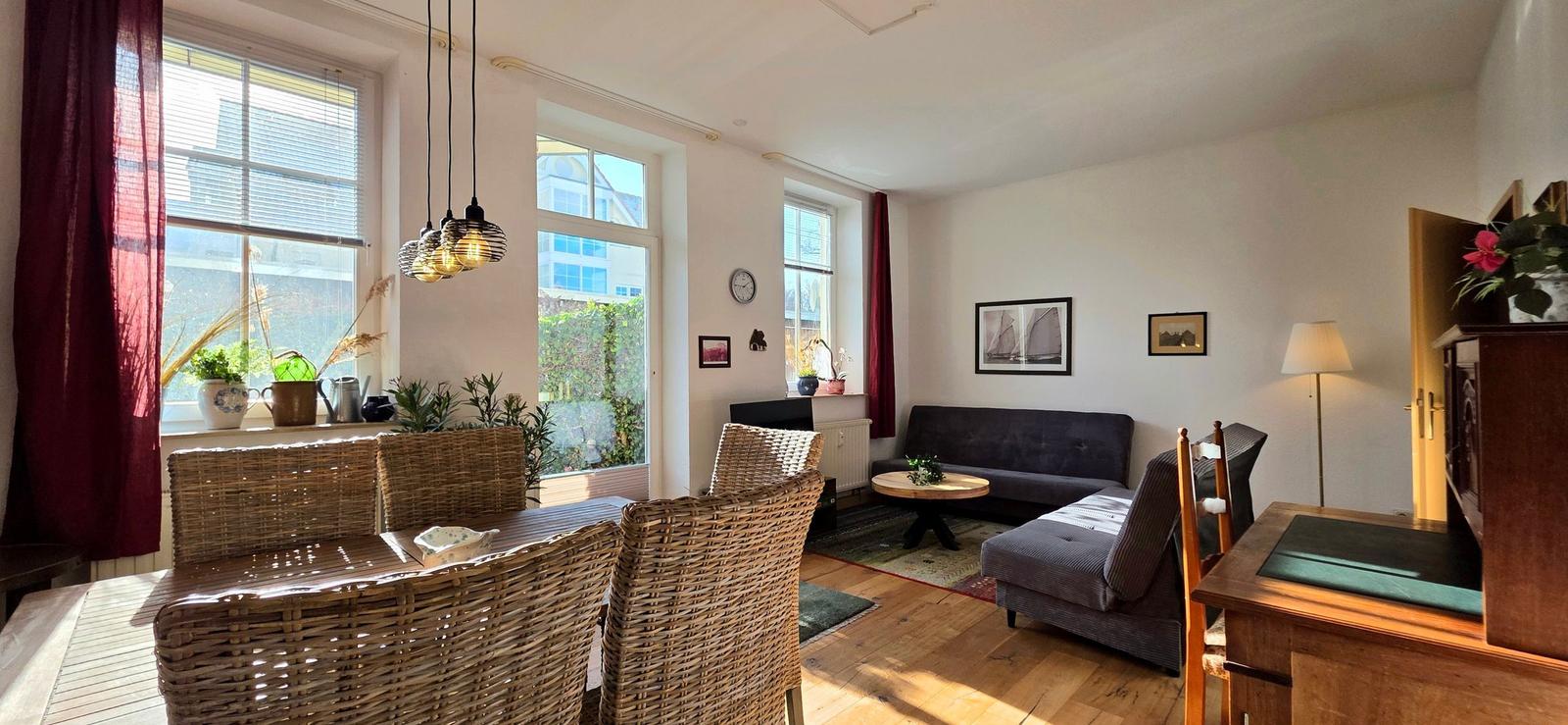 Living room with dining table, sofa, and windows. Wooden floor and plants.