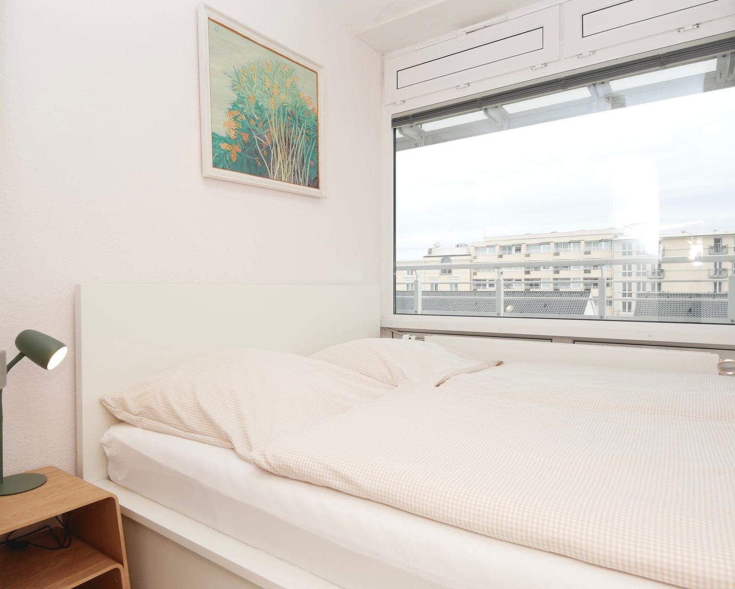 Schlafzimmer mit Bett, Nachttisch und Fenster mit Blick auf Gebäude.