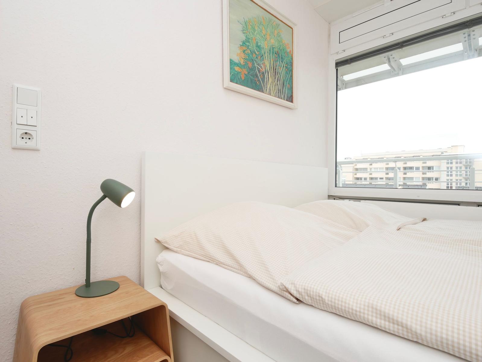 Schlafzimmer mit Bett, Nachttisch und Lampe. Fenster zeigt Blick auf Gebäude.
