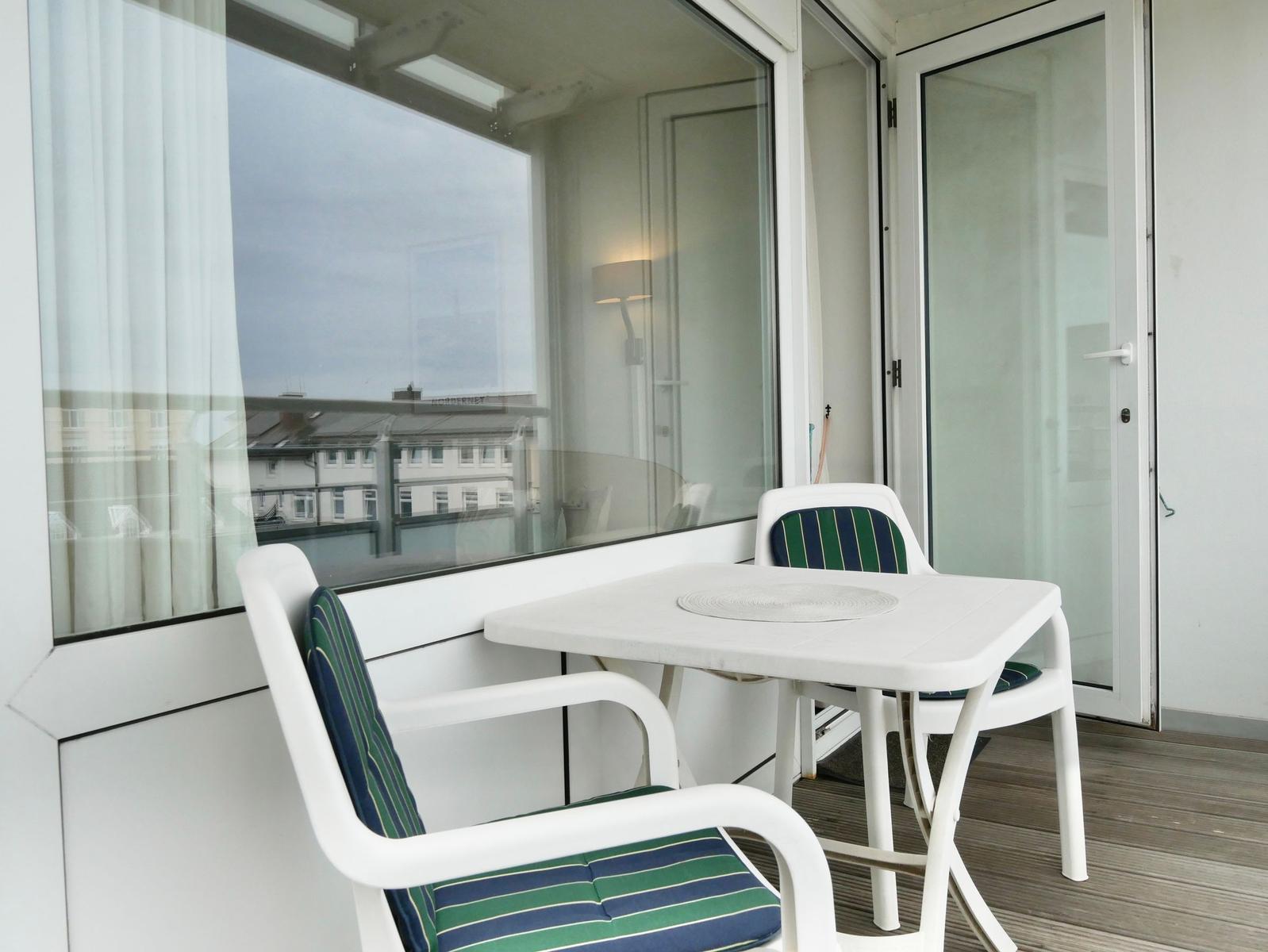 Terrasse mit Tisch und zwei Stühlen, Holzboden und Glaswand zur Wohnung.