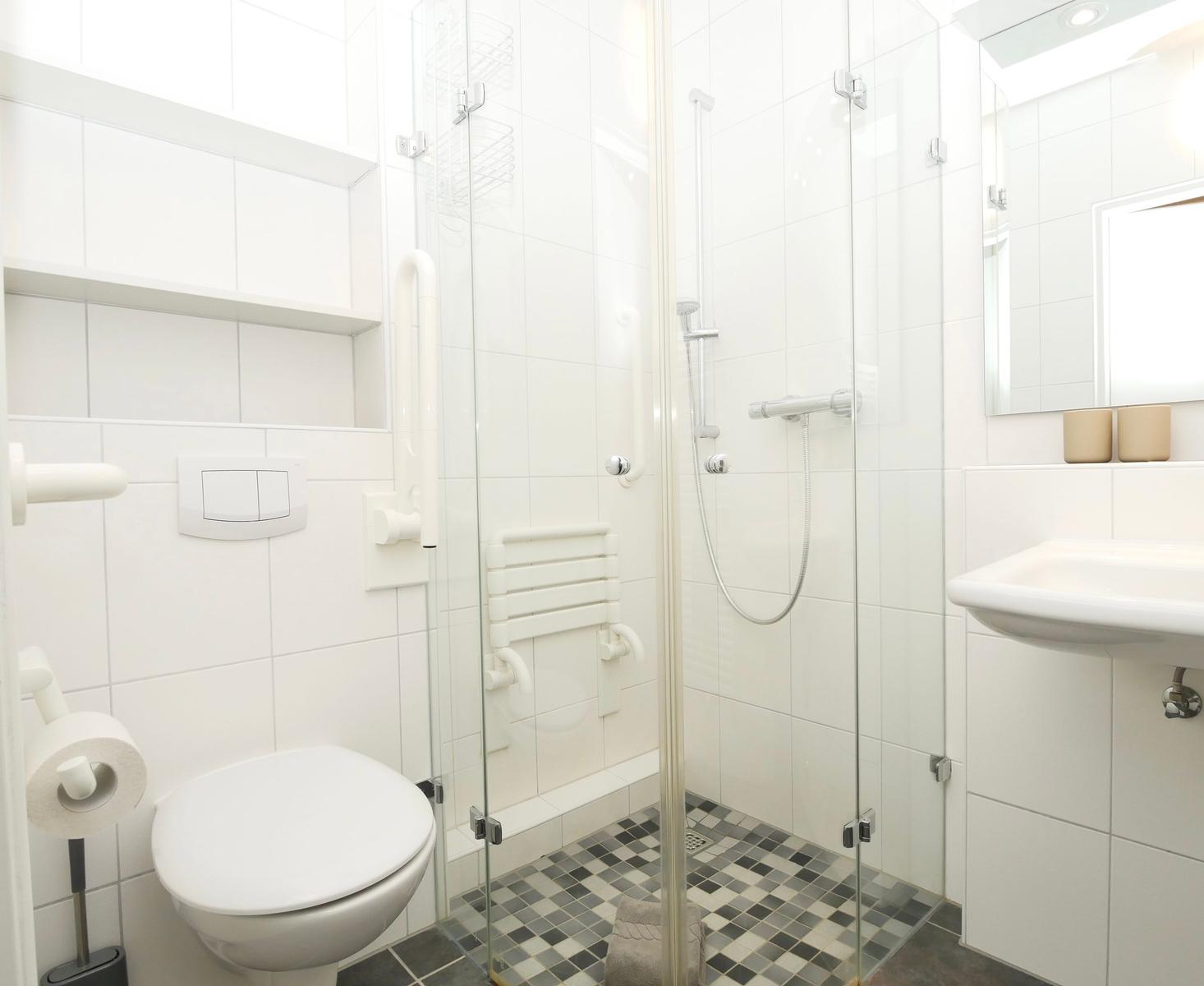 Badezimmer mit Dusche, Toilette und Waschbecken. Fliesenboden und weiße Wandfliesen.