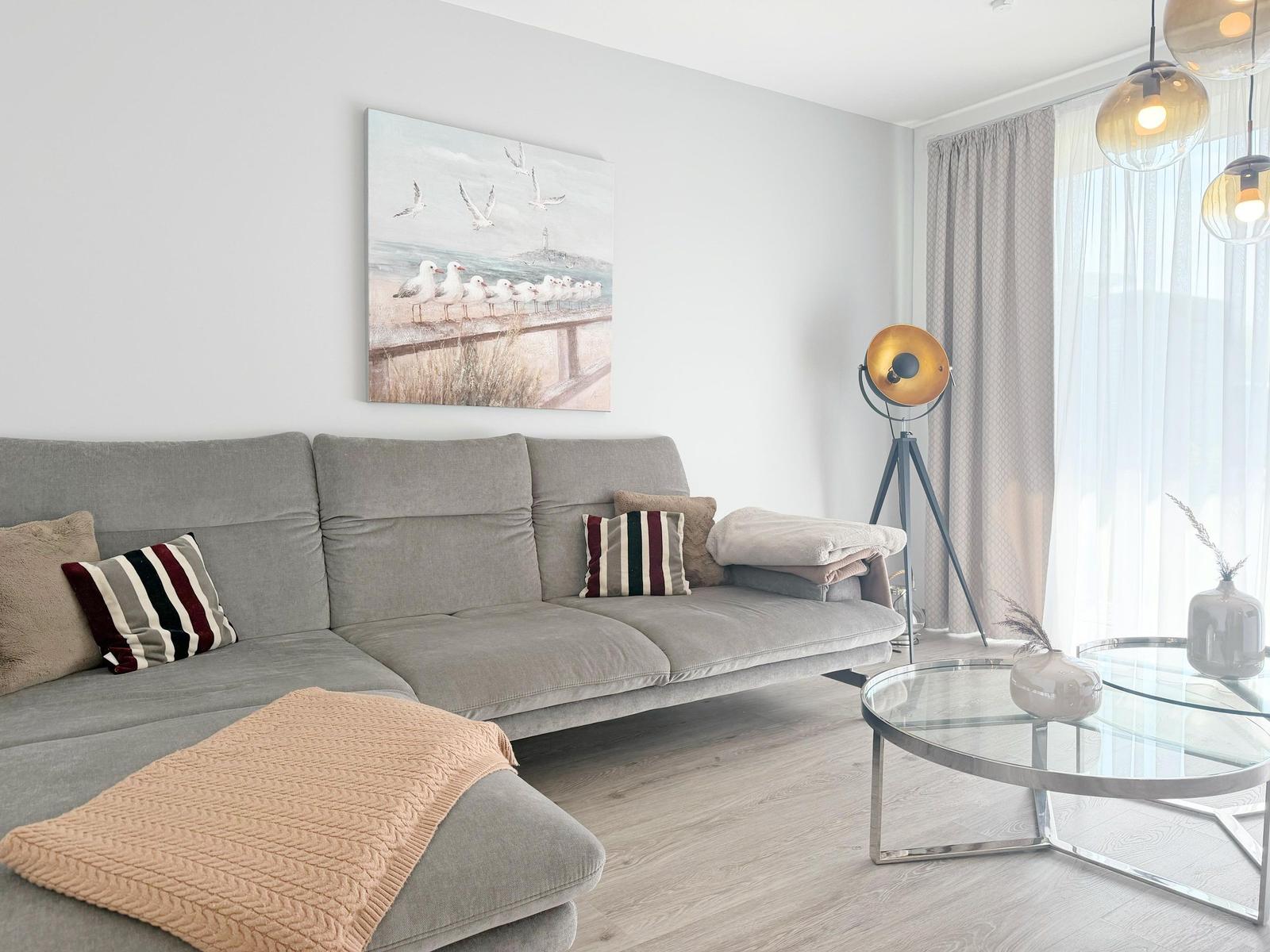 Ein Wohnzimmer mit grauem Sofa, Glas-Tisch und Wandbild mit Möwen.