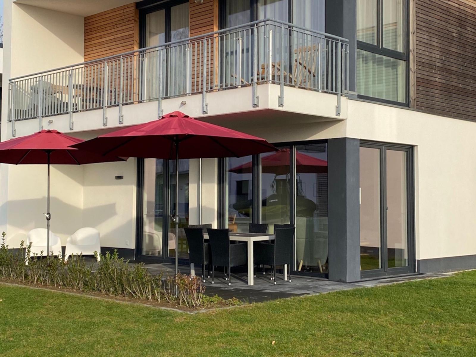 Terrasse mit Tisch, Stühlen und roten Sonnenschirmen vor modernem Haus mit Balkon.