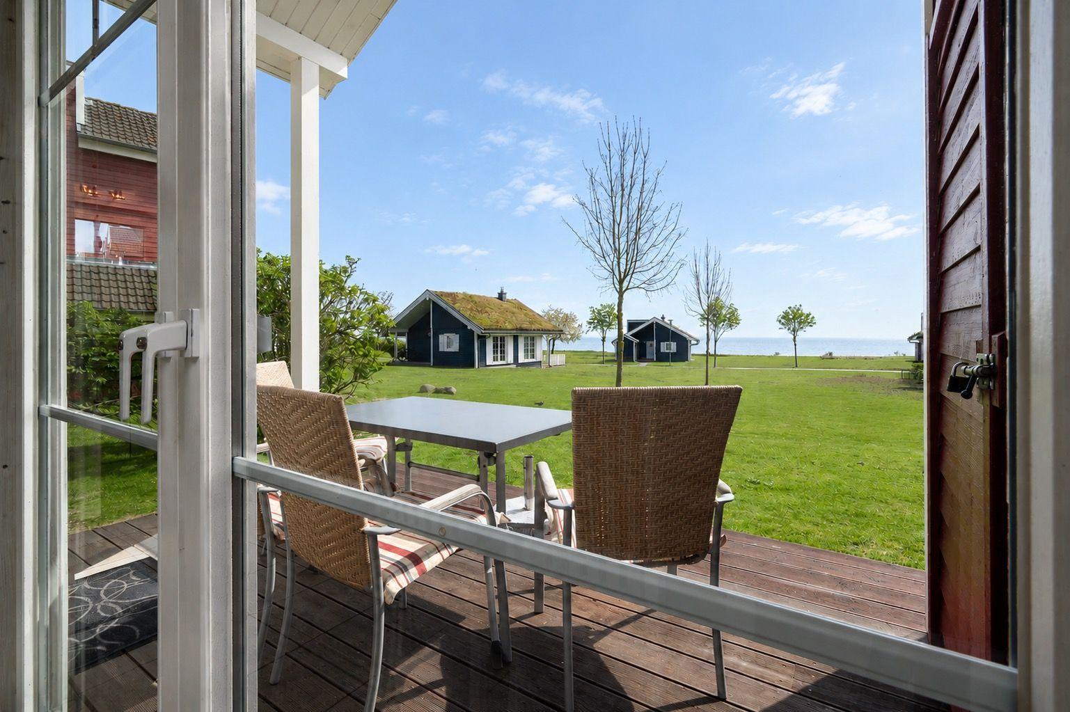 Terrace with table and chairs. View of green grass, houses, and sea.