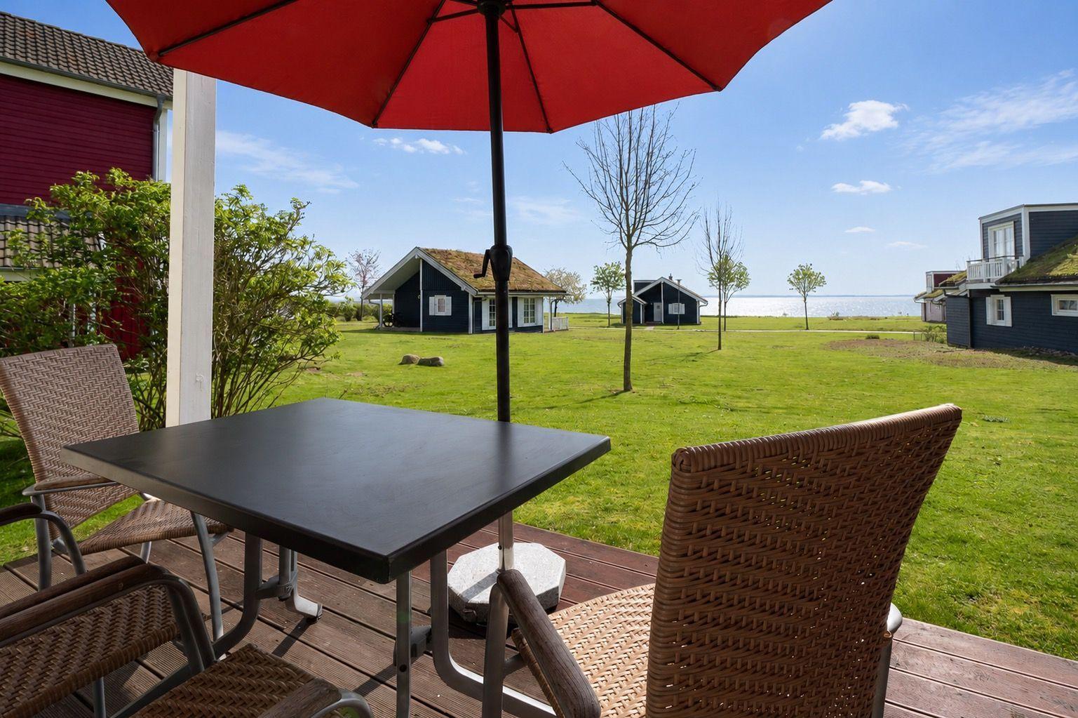 Terrace with table and chairs under a red umbrella. View of green grass and houses by the water.