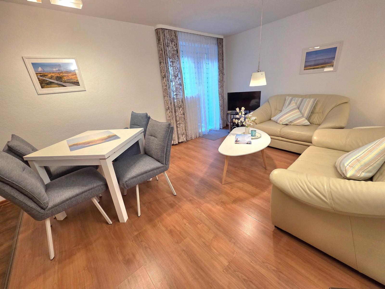 Wohn- und Essbereich mit Sofa, Tisch und Stühlen. Holzboden und Fenster mit Vorhängen.