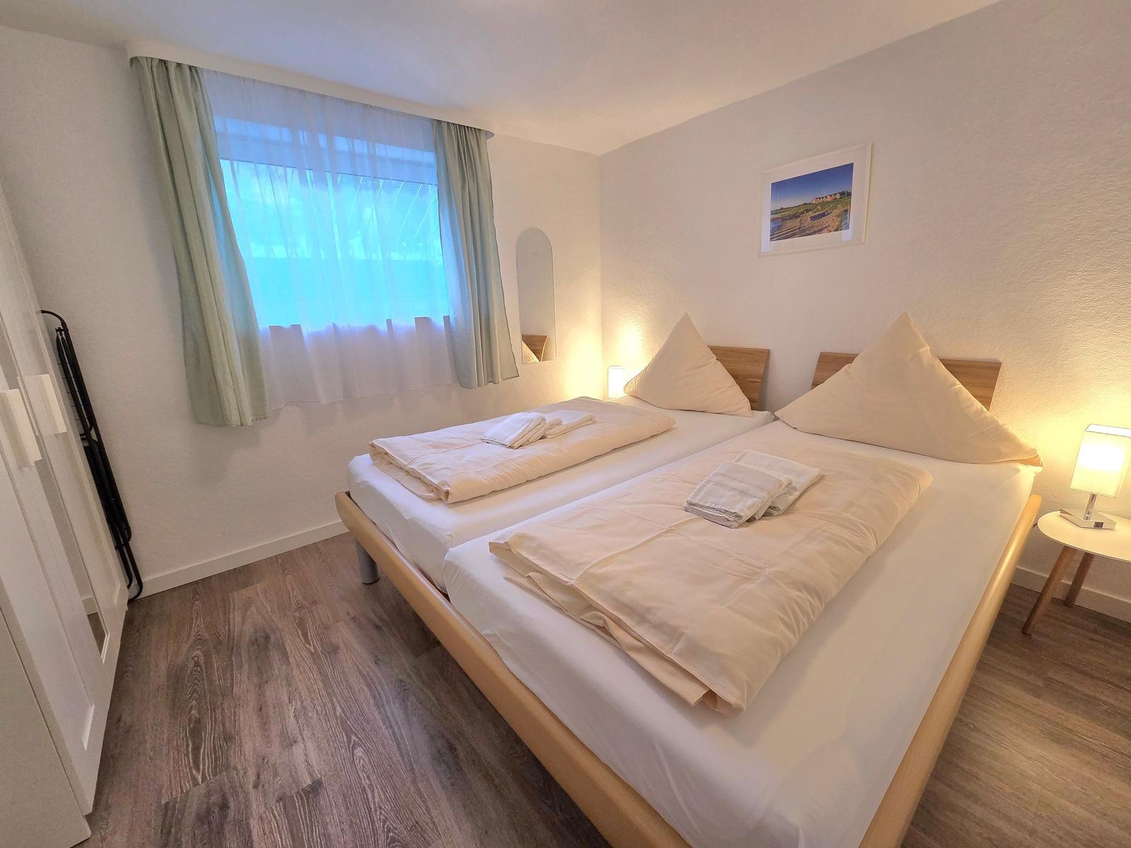 Zimmer mit Doppelbett, Fenster, Nachttisch und Wandbild.