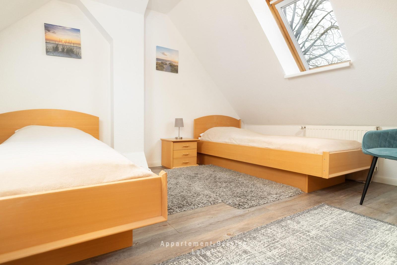 Zwei Betten, Nachttisch, Fenster und Teppiche in einem Dachzimmer.
