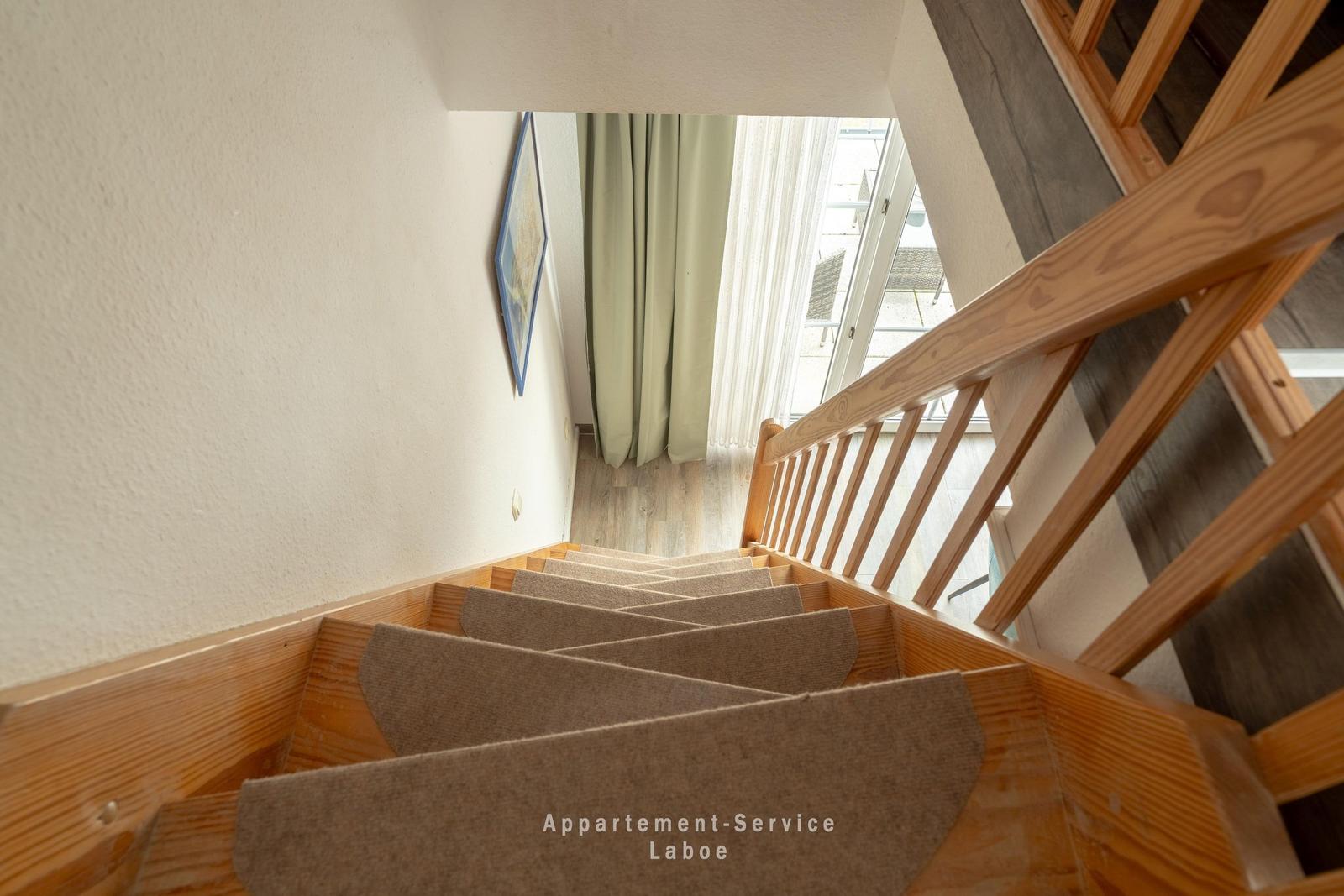 Holztreppe mit Teppichstufen führt zur Wohnung. Fenster mit Vorhängen und Terrasse sichtbar.