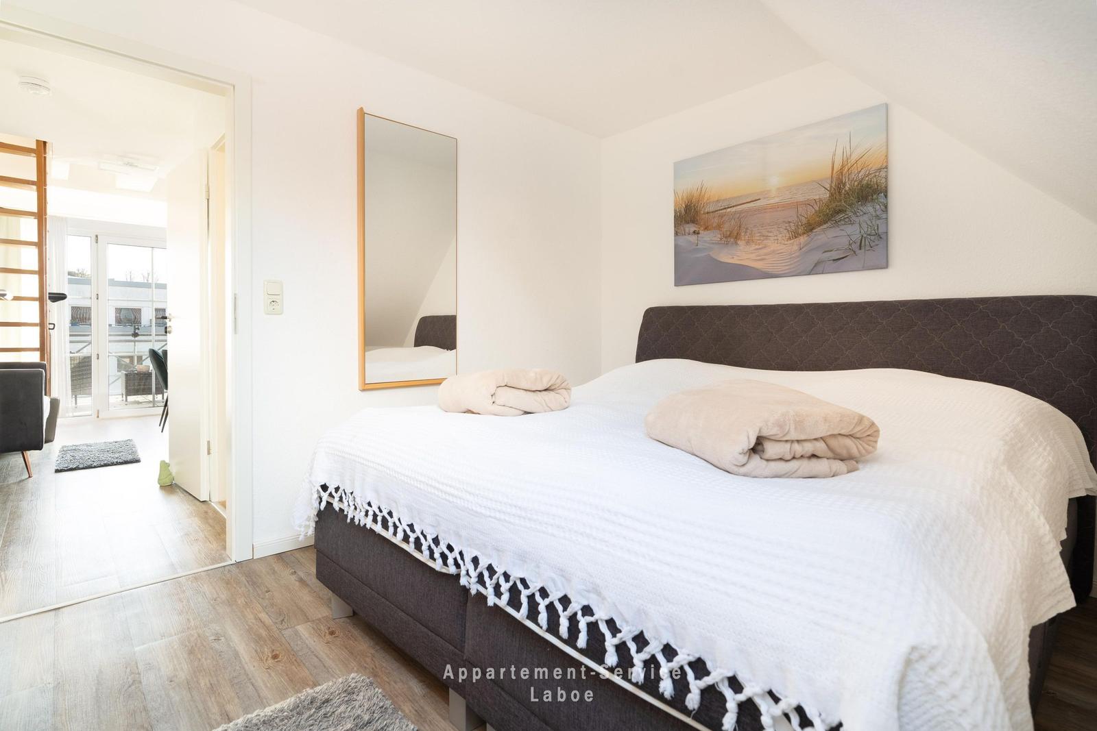 Schlafzimmer mit Bett, Wandbild und Spiegel. Tür führt in Wohnbereich mit Balkon.