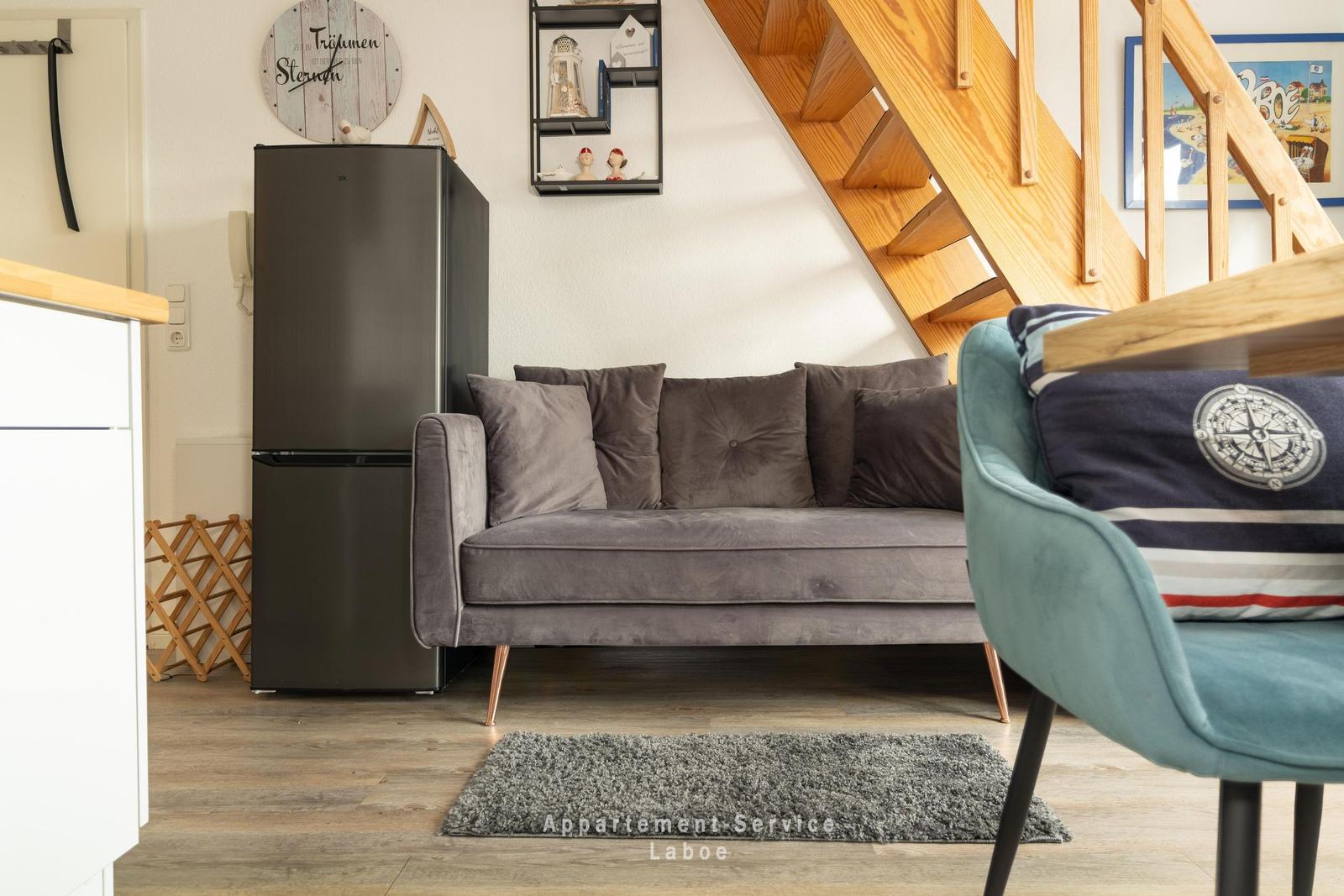 Ein grauer Sofa mit Kissen steht unter einer Holztreppe. Daneben steht ein schwarzer Kühlschrank.