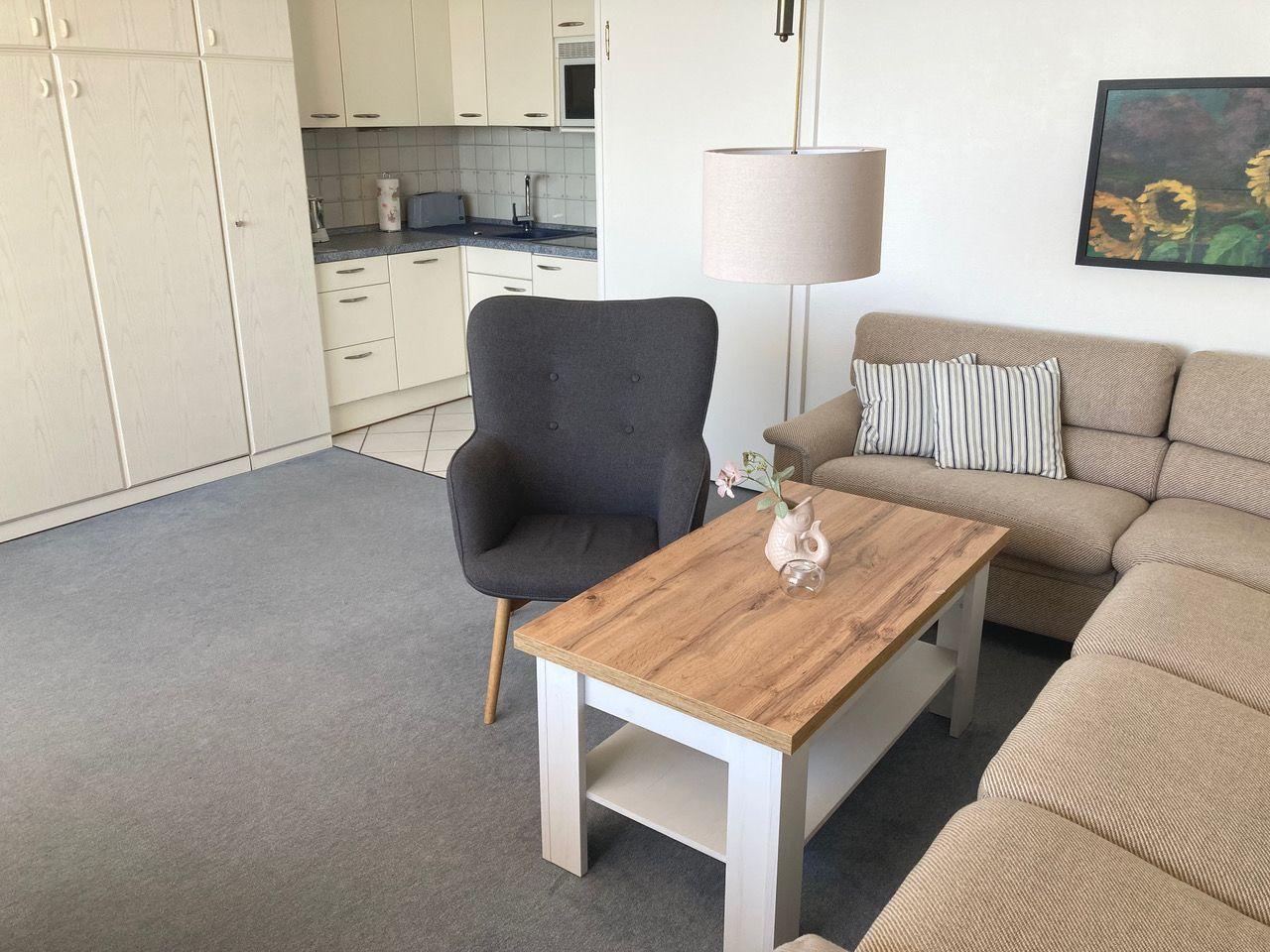 Living area with sofa, armchair, and table. Kitchen in the background.