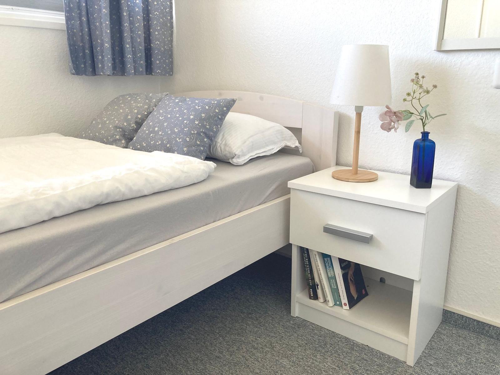 Bedroom with bed, nightstand, and lamp. Blue curtains and vase with flowers.