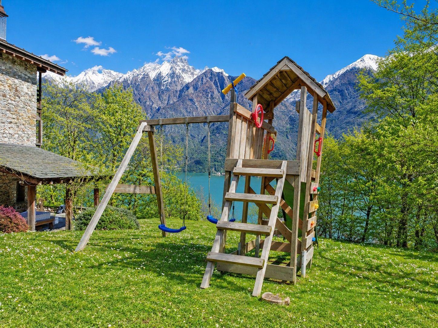 Wooden playground with swing and climbing wall in front of mountain lake and snow-capped peaks.