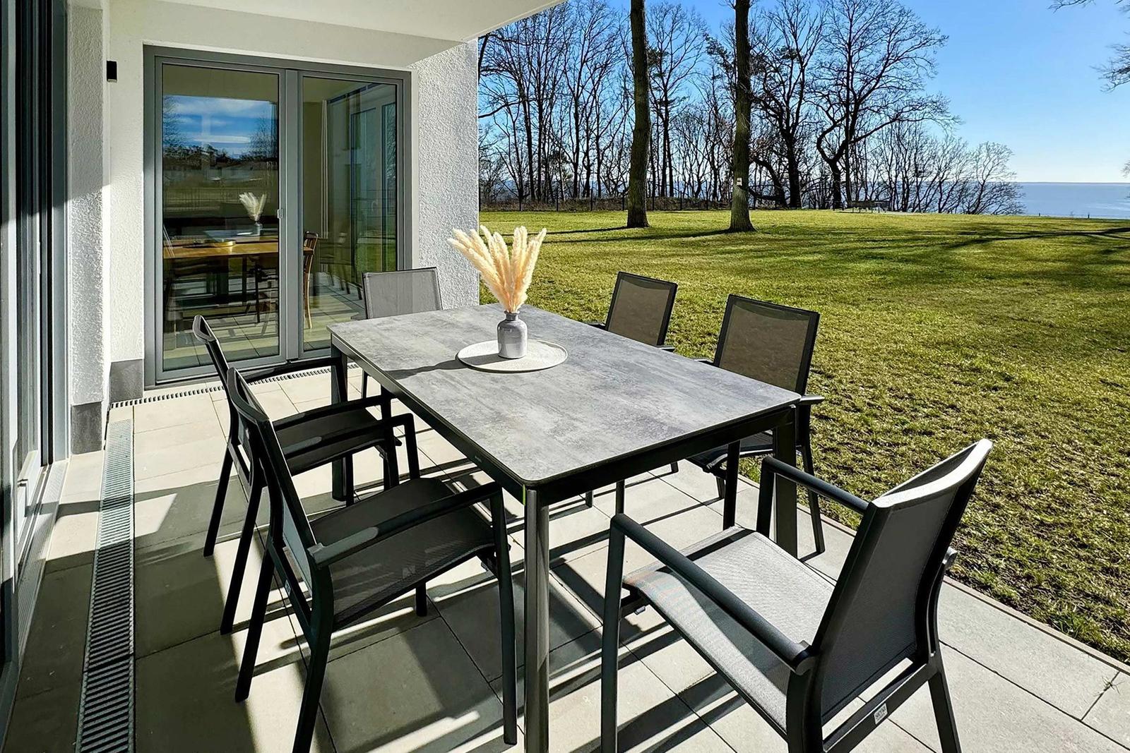Terrasse mit Esstisch und sechs Stühlen. Blick auf Grasfläche und Wasser.