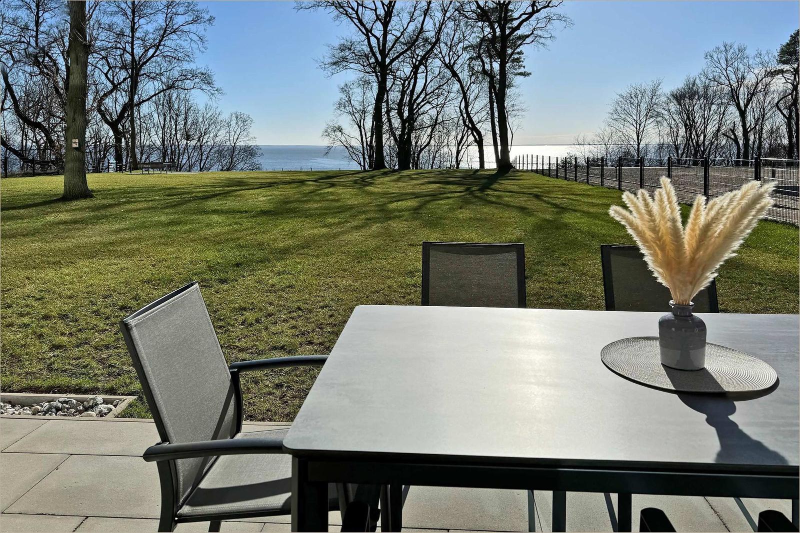 Terrasse mit Tisch und Stühlen, Blick auf Grasfläche und See.
