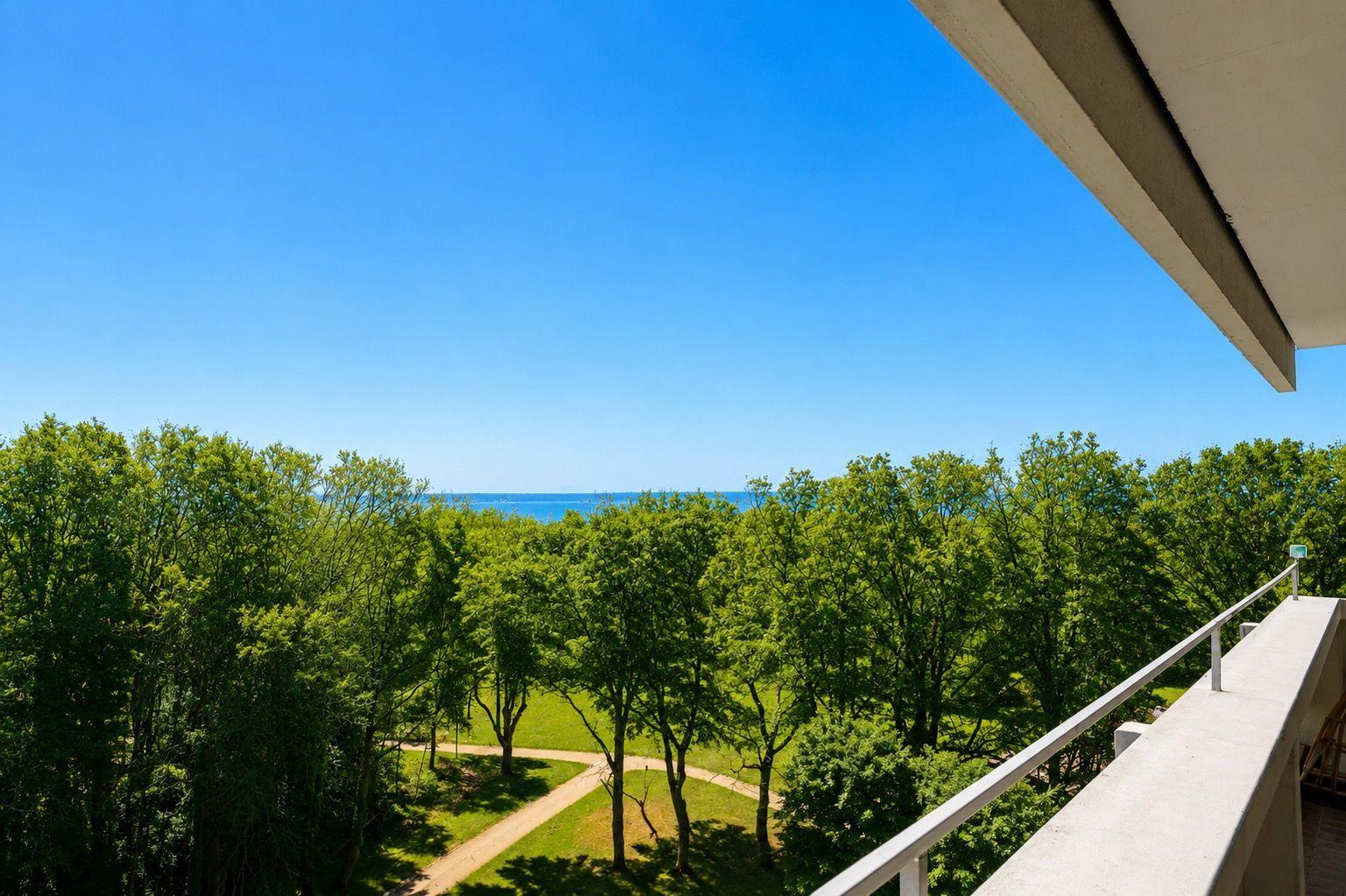Blick von Balkon auf grüne Bäume und blaues Meer unter klarem Himmel