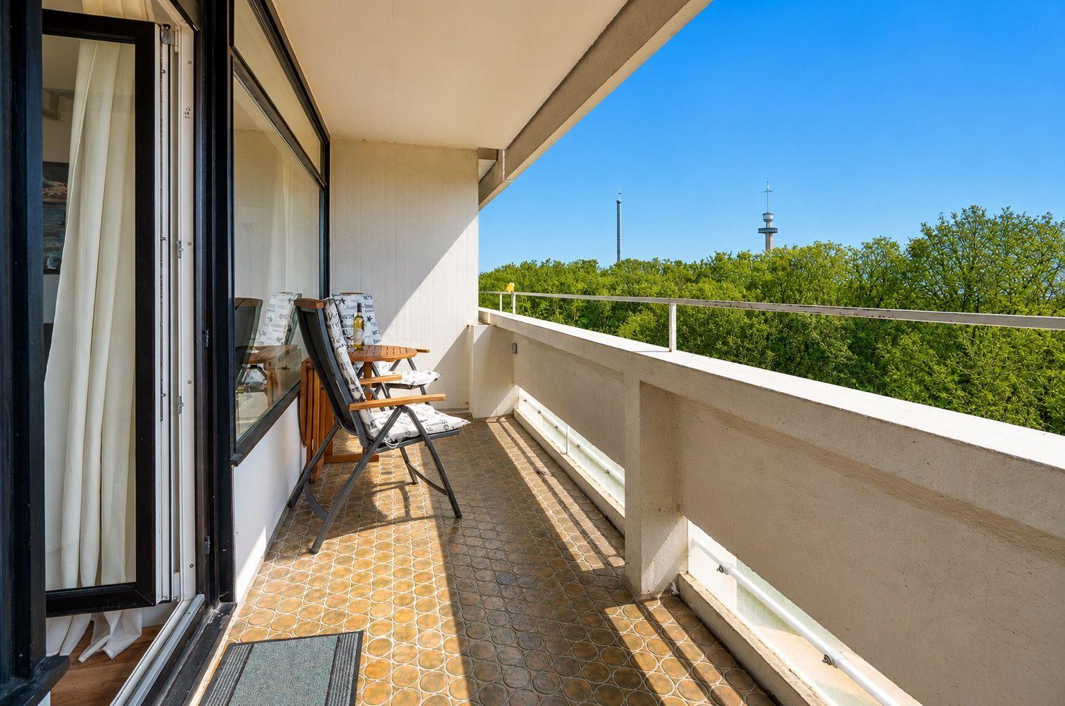 Terrasse mit Sitzgelegenheit und Blick auf grüne Bäume und einen Turm.