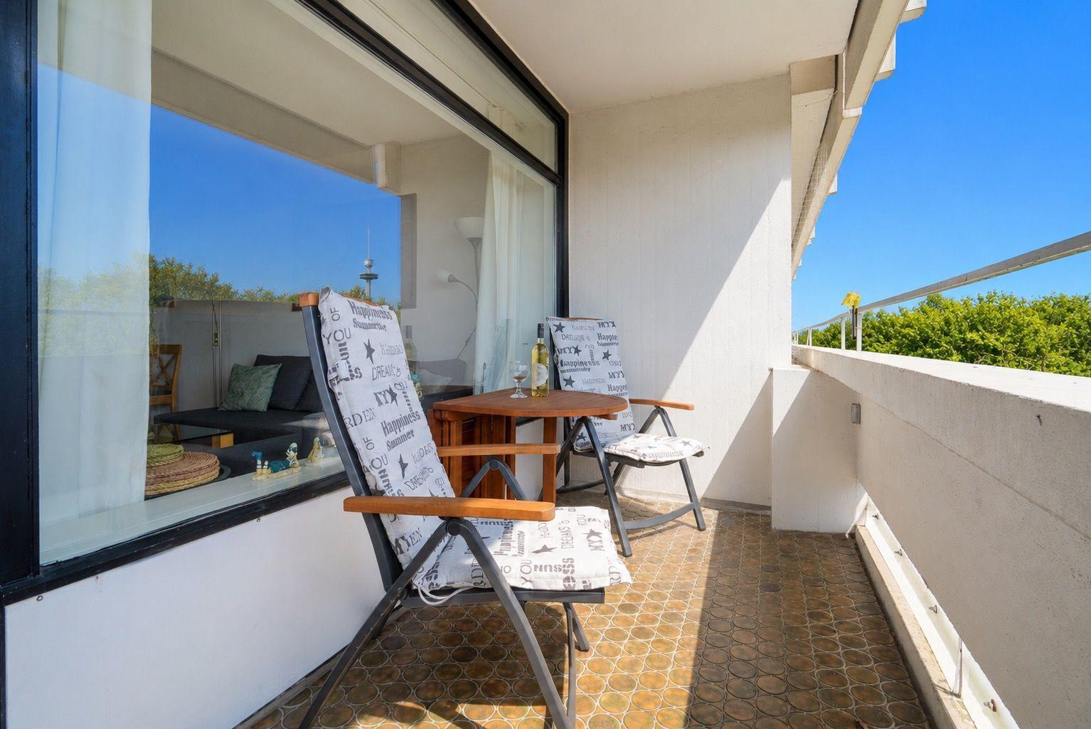 Terrasse mit zwei Stühlen, Tisch und Flasche. Blick auf grüne Bäume und blauen Himmel.