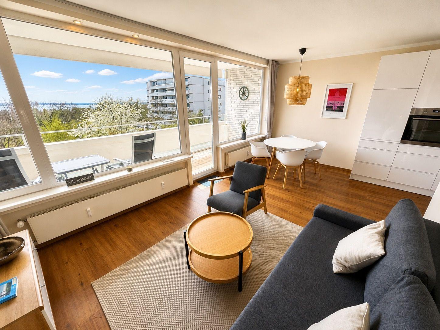 Living area with sofa, table, and chair. Large windows with view of balcony and city.