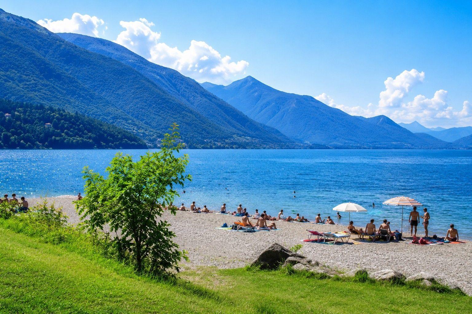 Strand mit Menschen, blauem Wasser und Bergen im Hintergrund.