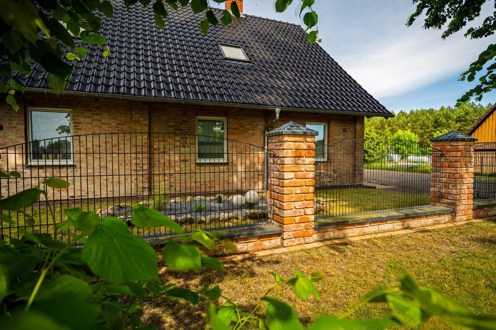 Haus mit Ziegelfassade und Dachgaube hinter Zaun mit Steinpfeilern.