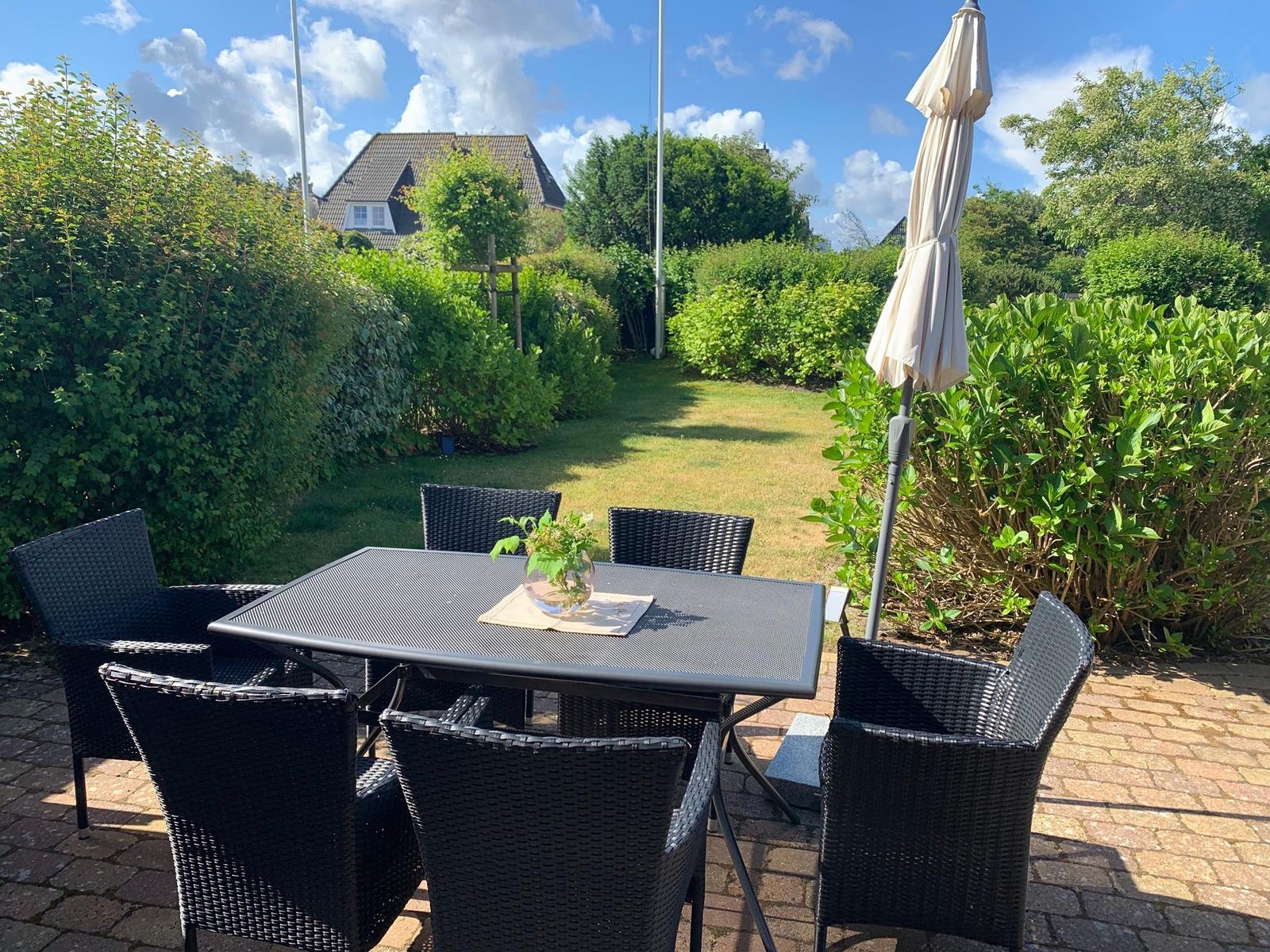 Terrasse mit Tisch und sechs Stühlen, Sonnenschirm, Grünflächen und Gartenhaus im Hintergrund.