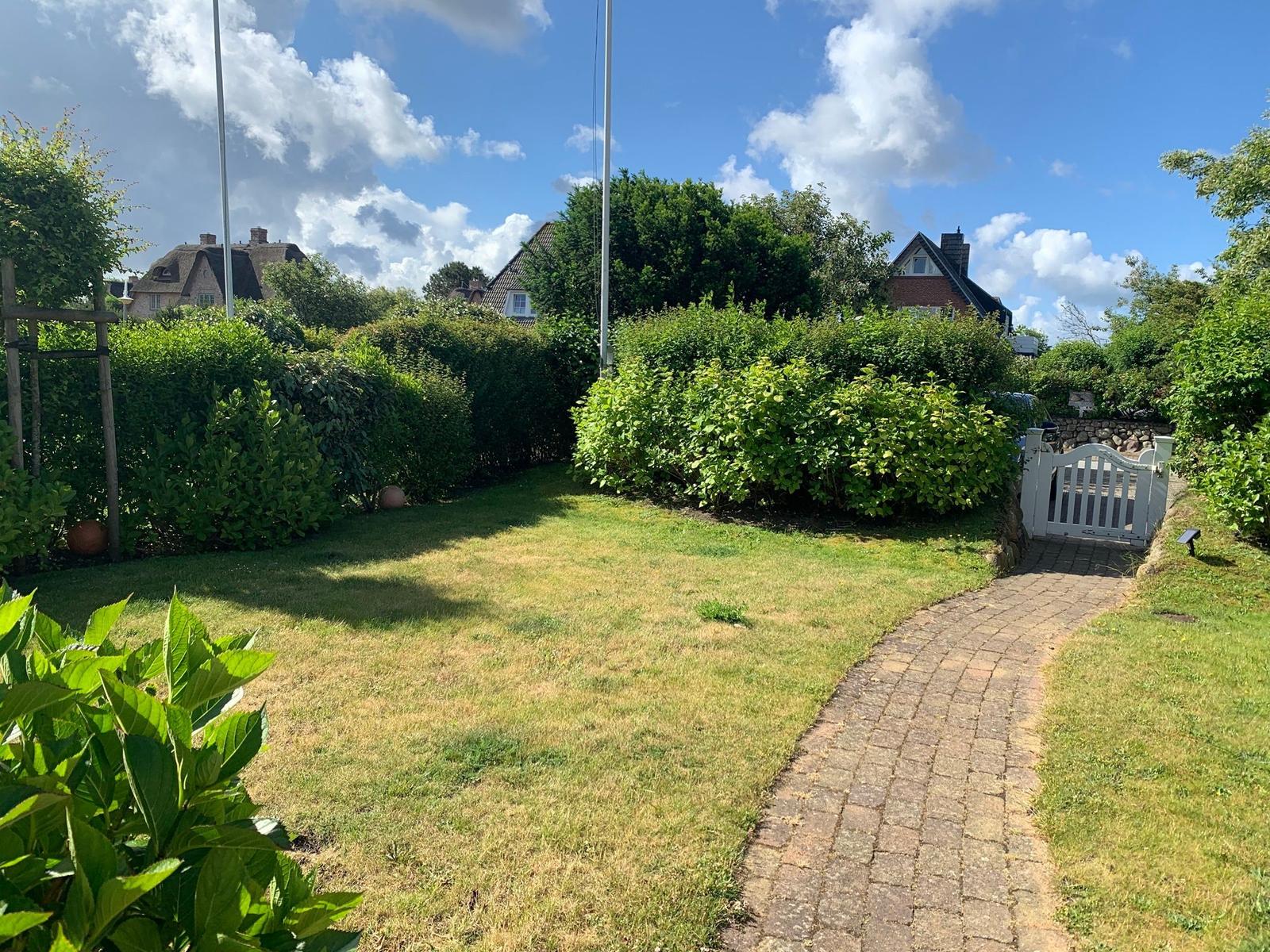 Ein gepflegter Garten mit Rasen, Hecken und einem weißen Tor. Im Hintergrund sind Häuser und Bäume zu sehen.