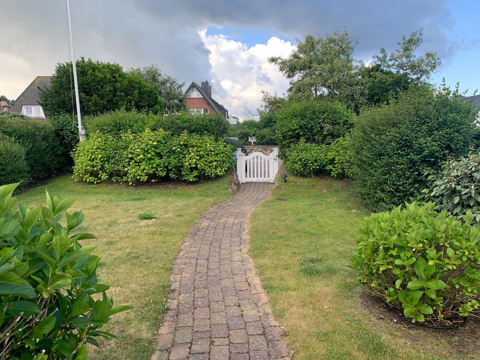 Ein gepflasterter Weg führt durch einen grünen Garten zu einem weißen Tor.