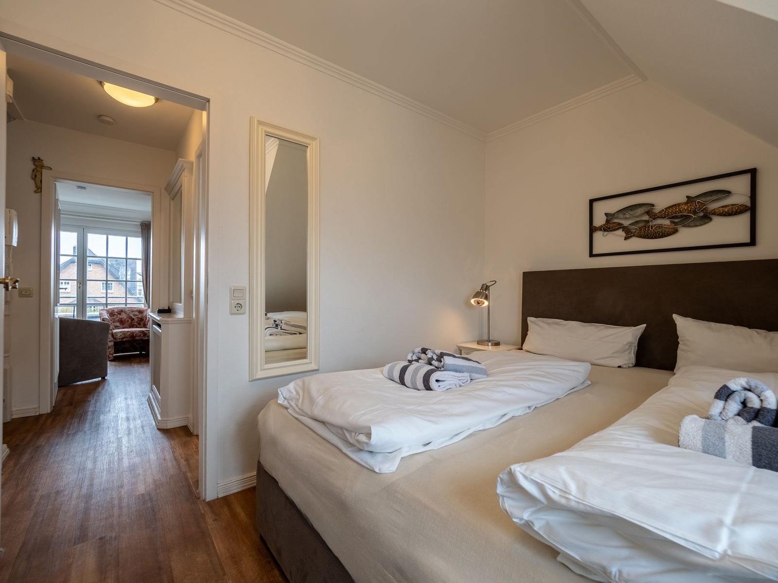 Zwei Betten mit weißen Laken, Wandspiegel und Bild an der Wand. Durchgang zu Wohnbereich mit Blick auf Balkon.