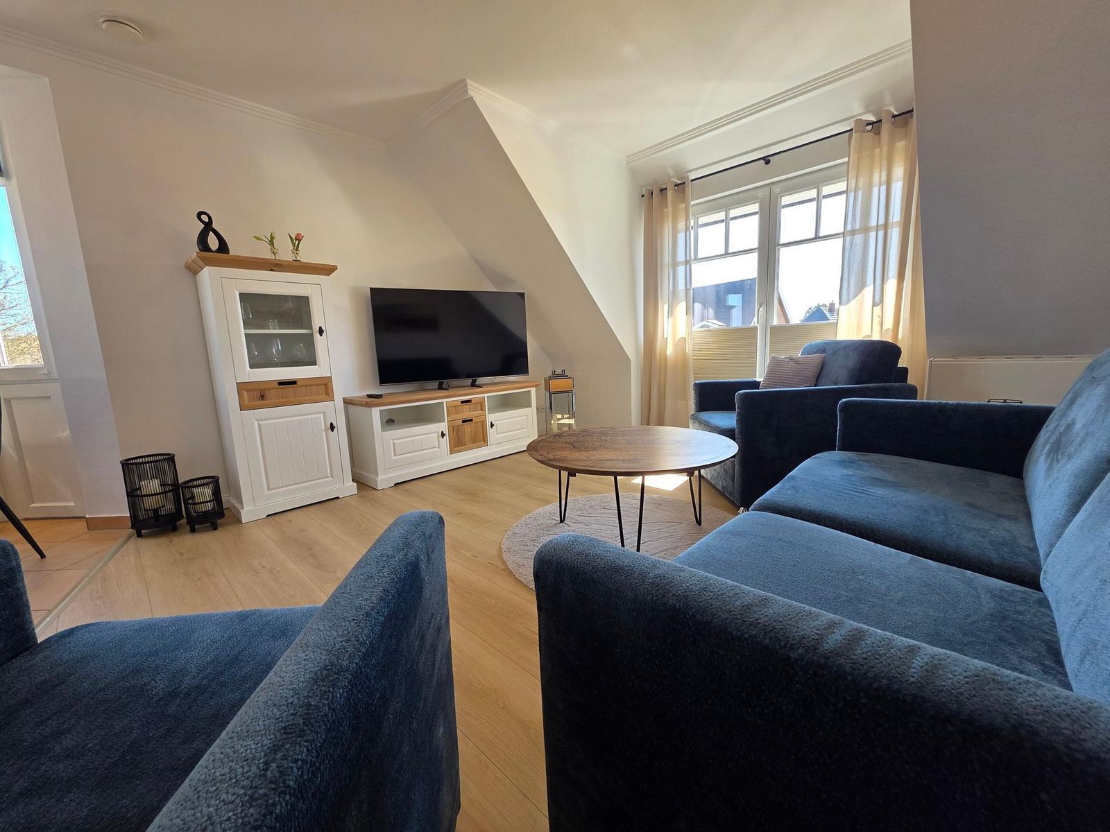 Wohnzimmer mit blauem Sofa, TV und Holzboden. Fenster mit Vorhängen.