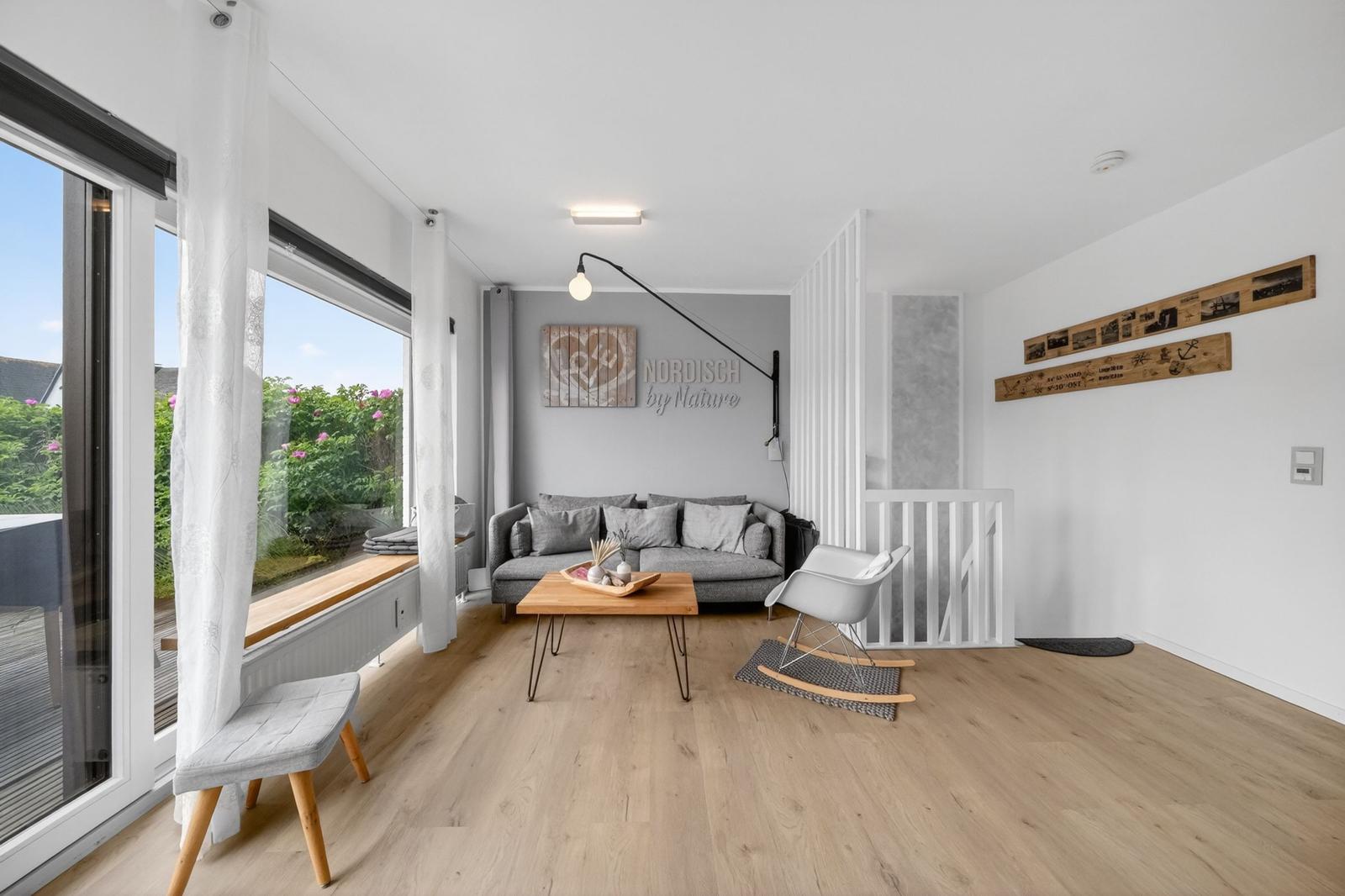 Wohnzimmer mit Sofa, Tisch und Sessel. Große Fenster und Holzboden.