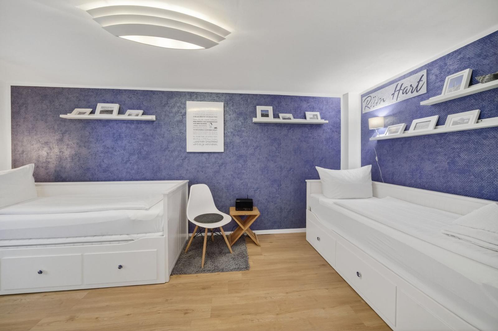 Zimmer mit zwei weißen Betten, blauer Wand und weißen Regalen.