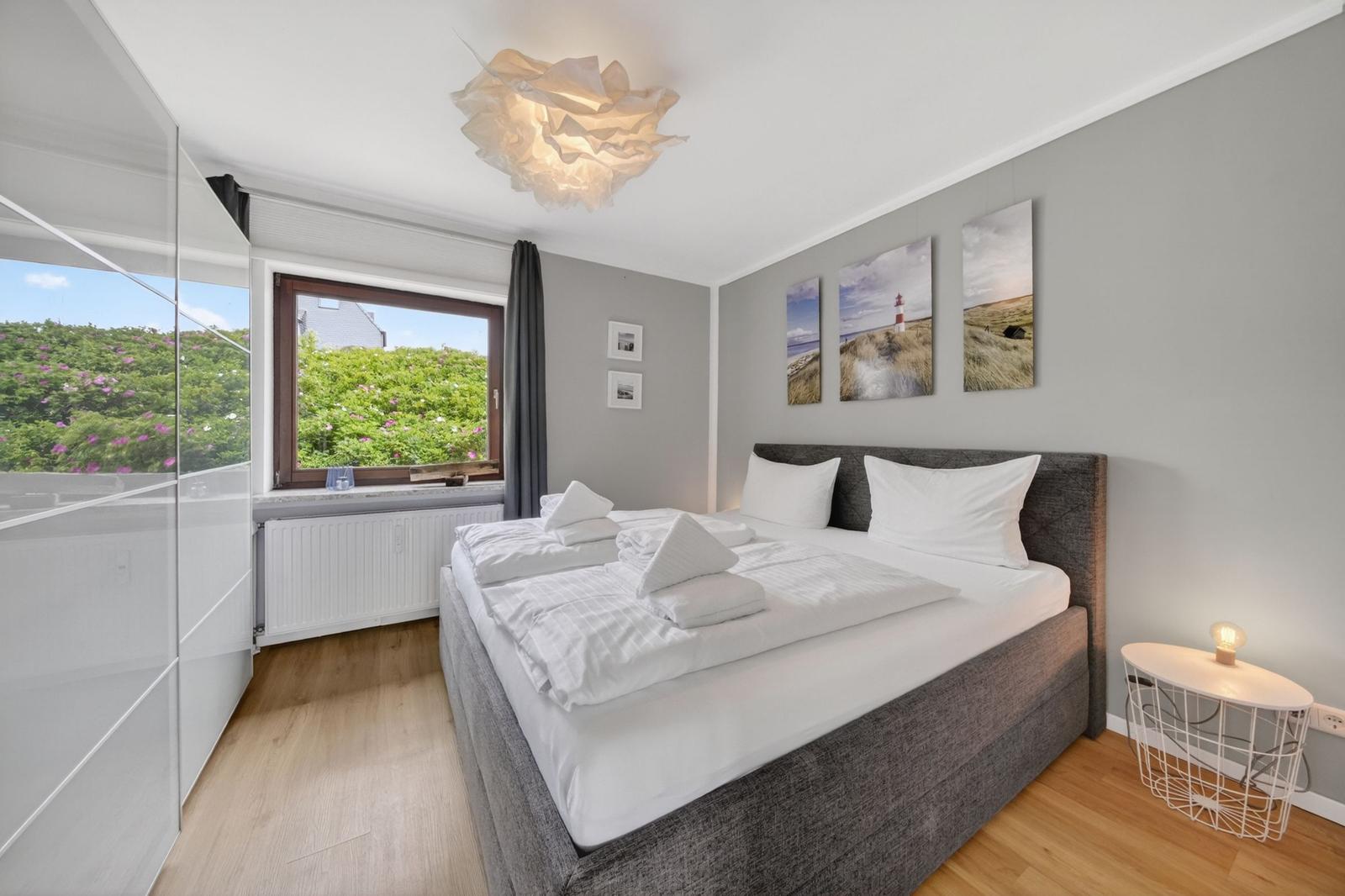 Schlafzimmer mit Doppelbett, Fenster mit Blick auf Grünfläche und Wandbildern mit Leuchtturm.