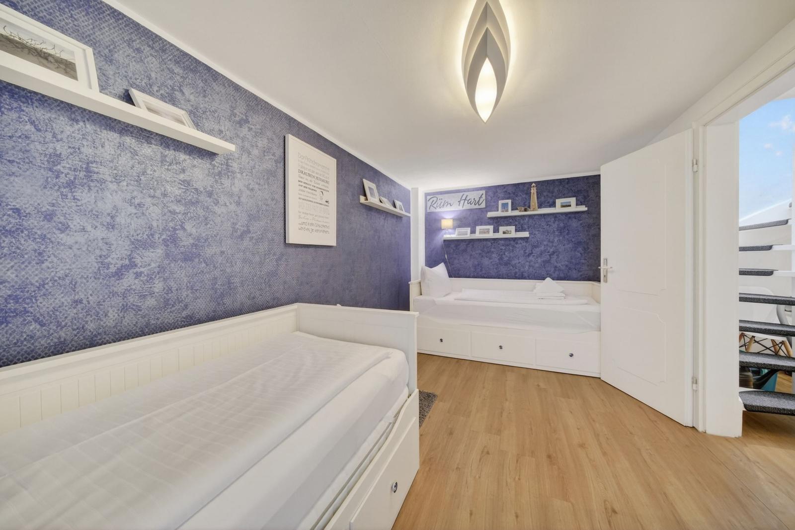 Zwei weiße Betten mit Schubladen, lila Wand, Holzboden und Deckenleuchte.