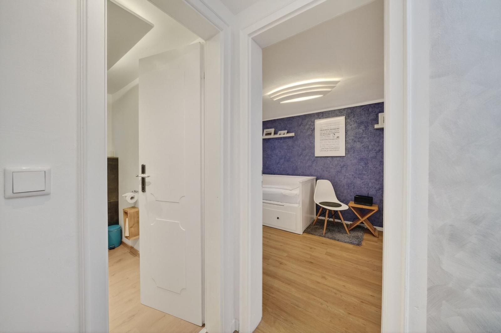 Schlafzimmer mit blauer Wand und weißem Bett. Nebenraum mit Toilette und Tür.
