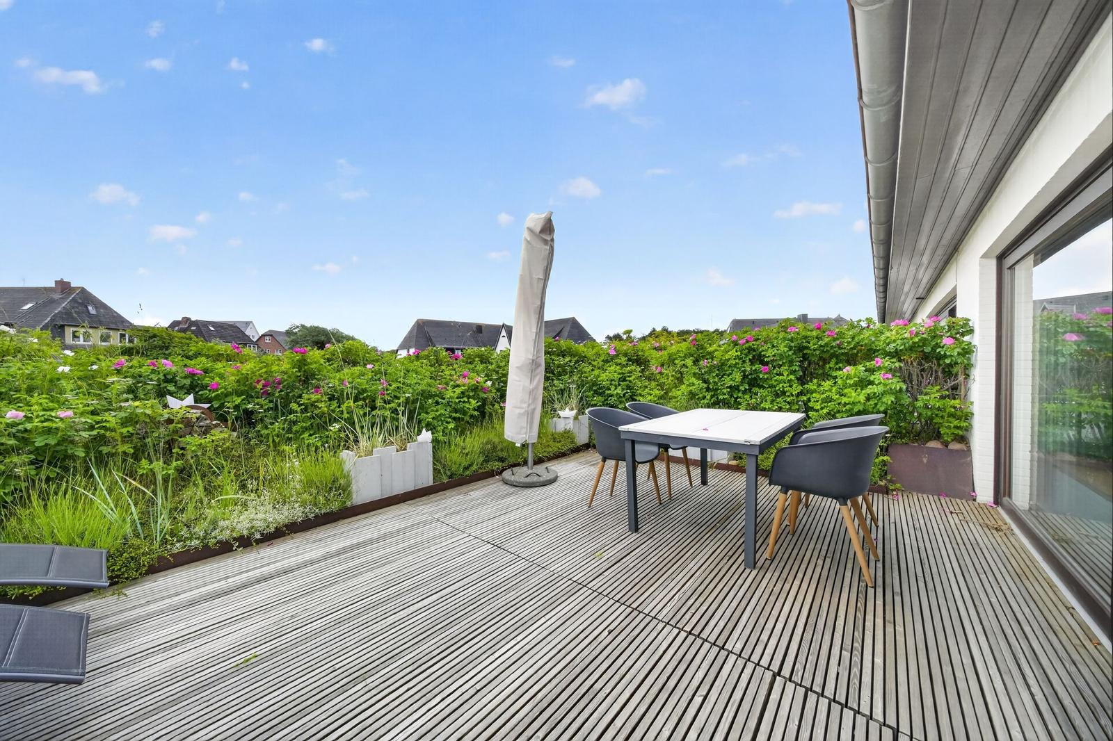 Terrasse mit Tisch, Stühlen und Sonnenschirm. Grünfläche und Blick auf Wohnhäuser.