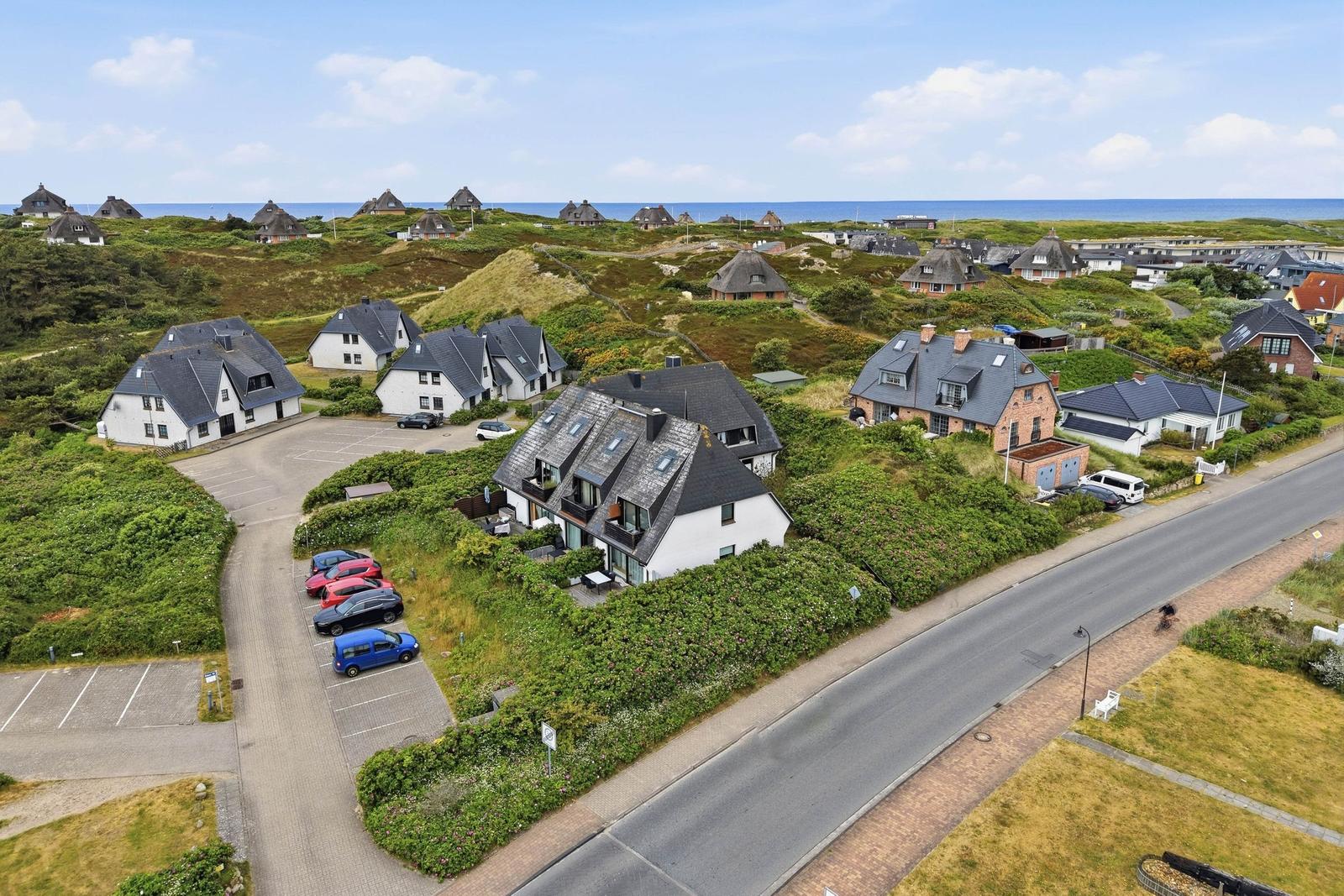 Haus mit Terrasse und Parkplatz, umgeben von Grünflächen und Blick aufs Meer.
