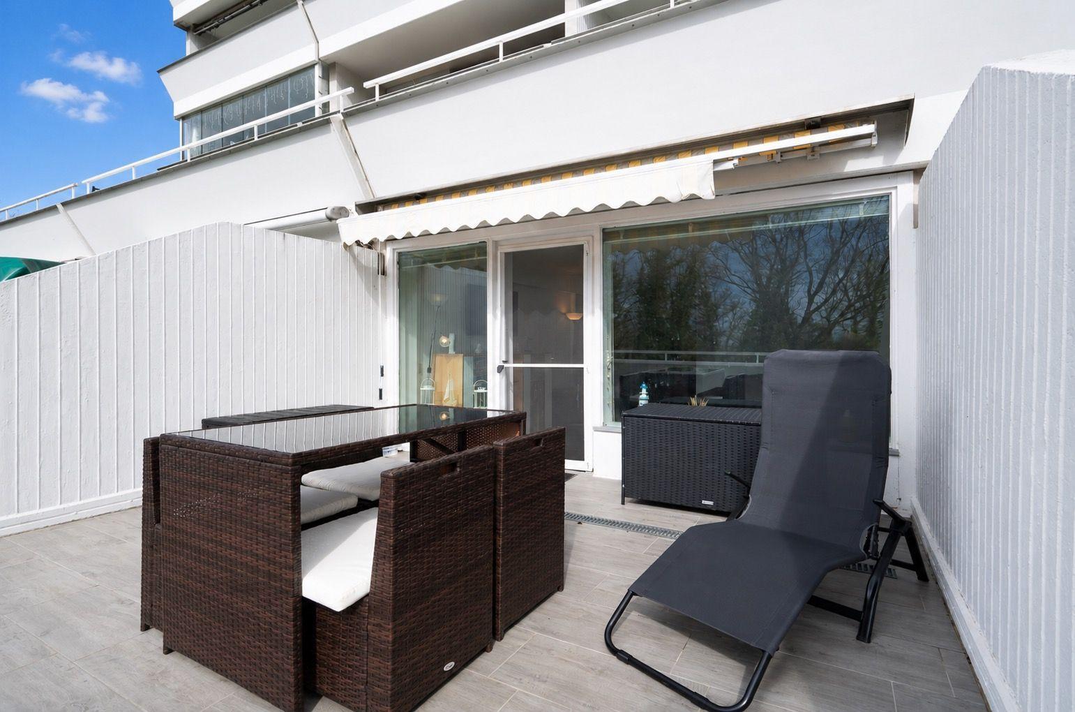 Terrace with lounge furniture, table, and seating. Background: building with windows and white walls.