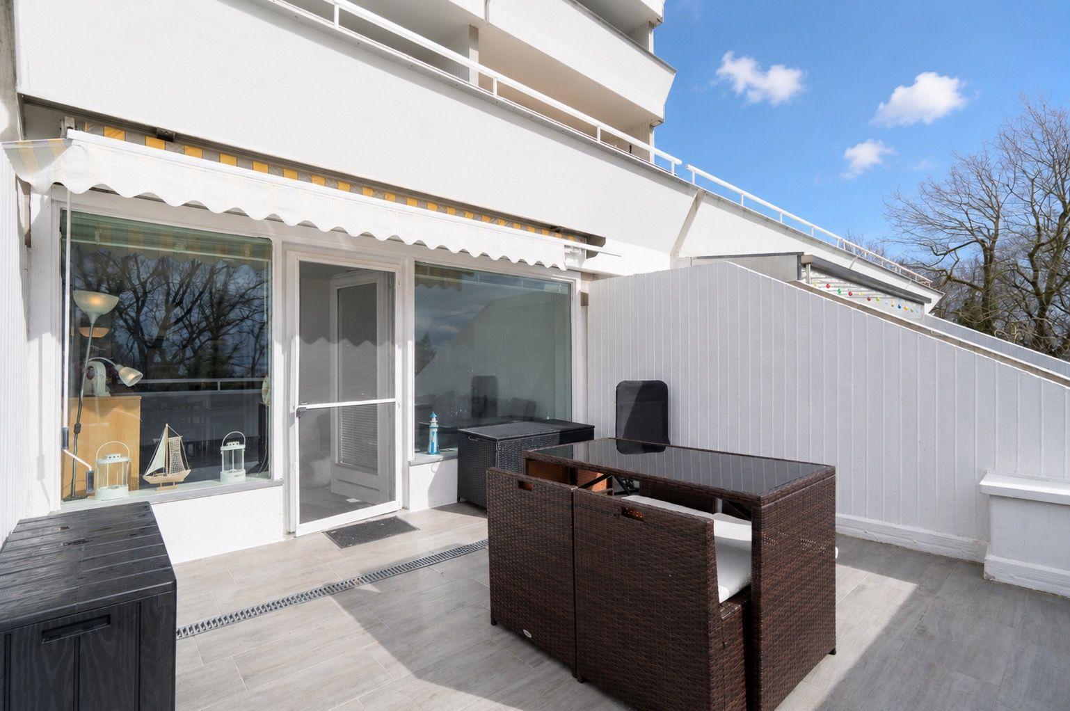 Terrace with seating area, glass front, and white wall. Sky is blue with white clouds.
