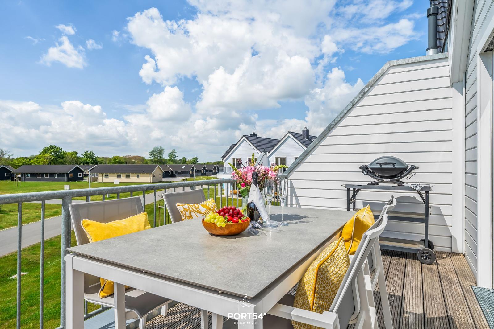 Terrasse mit Tisch, Stühlen und Grill. Blick auf Ferienhäuser und Grünflächen.