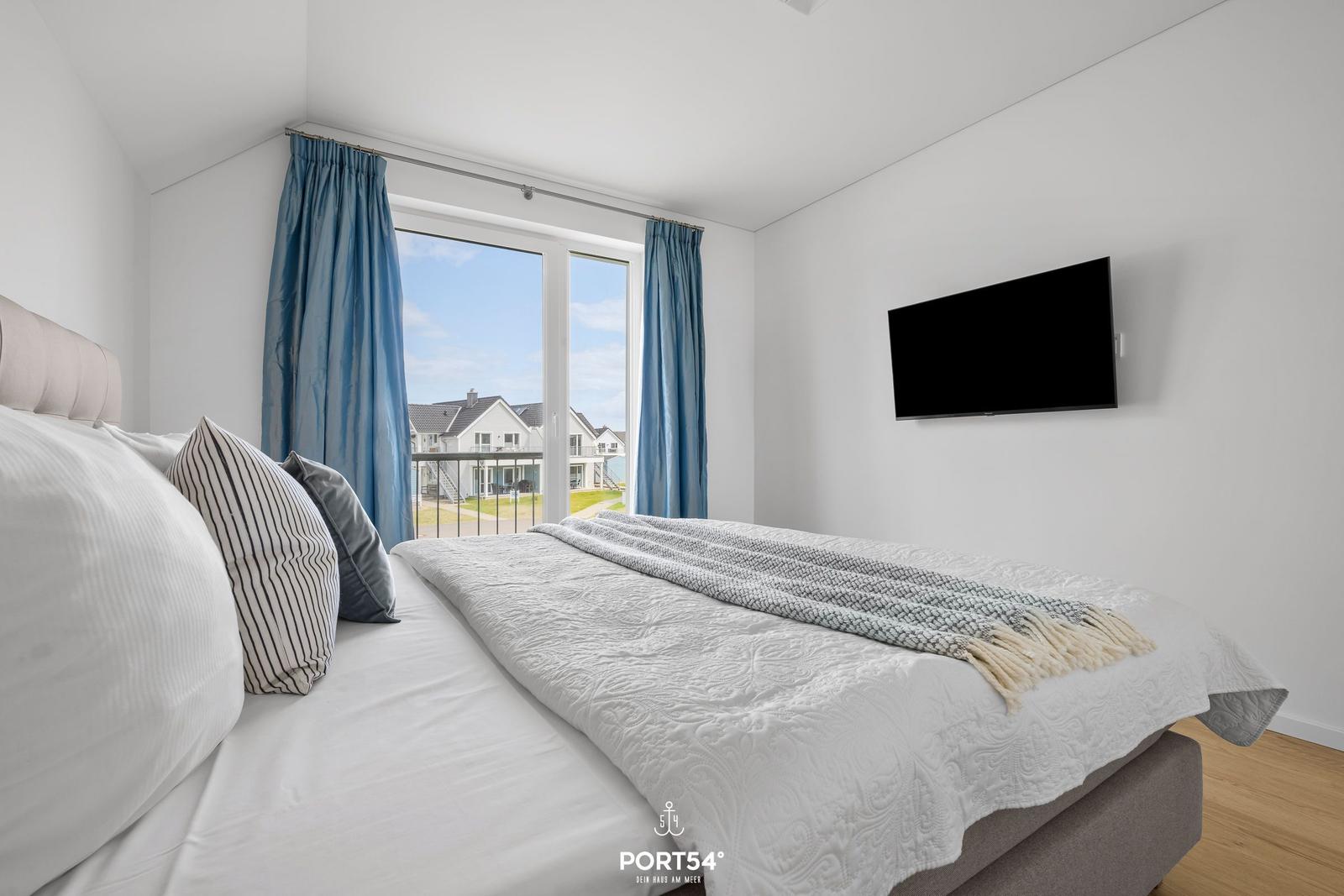 Schlafzimmer mit Blick auf Balkon und Strand. Bett, TV und blaue Vorhänge.