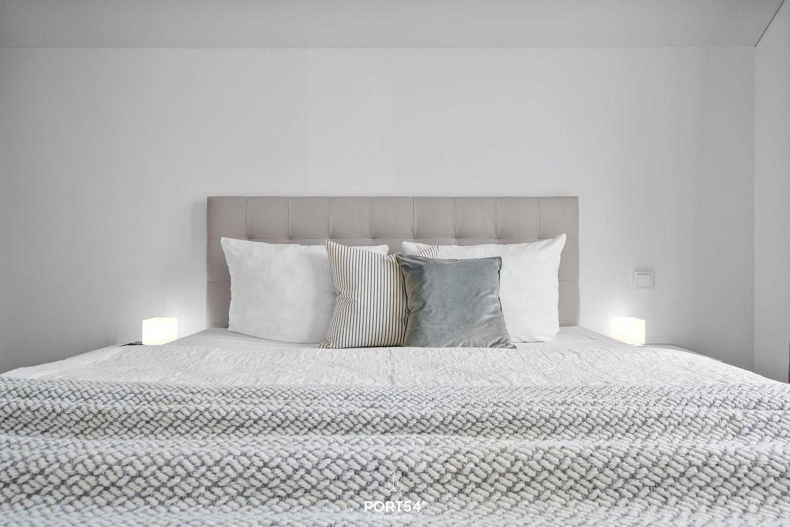 Doppelbett mit Kopfkissen und Decke. Zwei Nachttische mit Lampen.