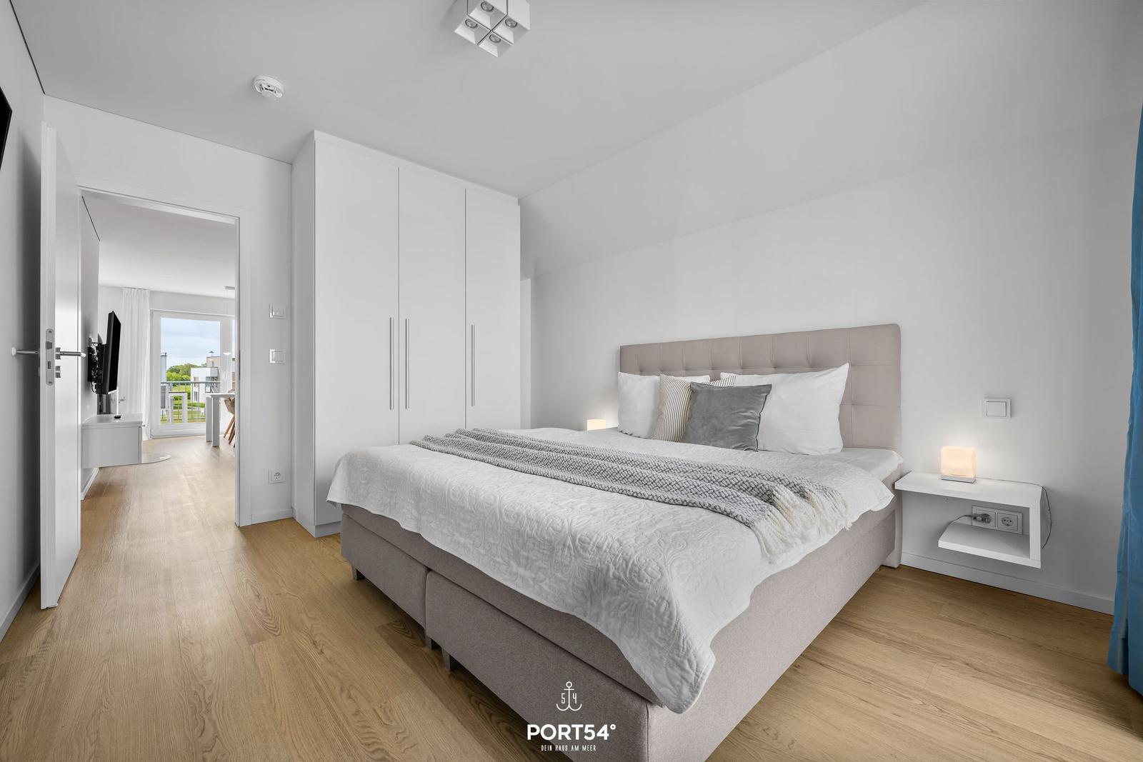 Schlafzimmer mit Bett, Nachttisch und Kleiderschrank. Durchgang zum Wohnbereich mit Balkon.