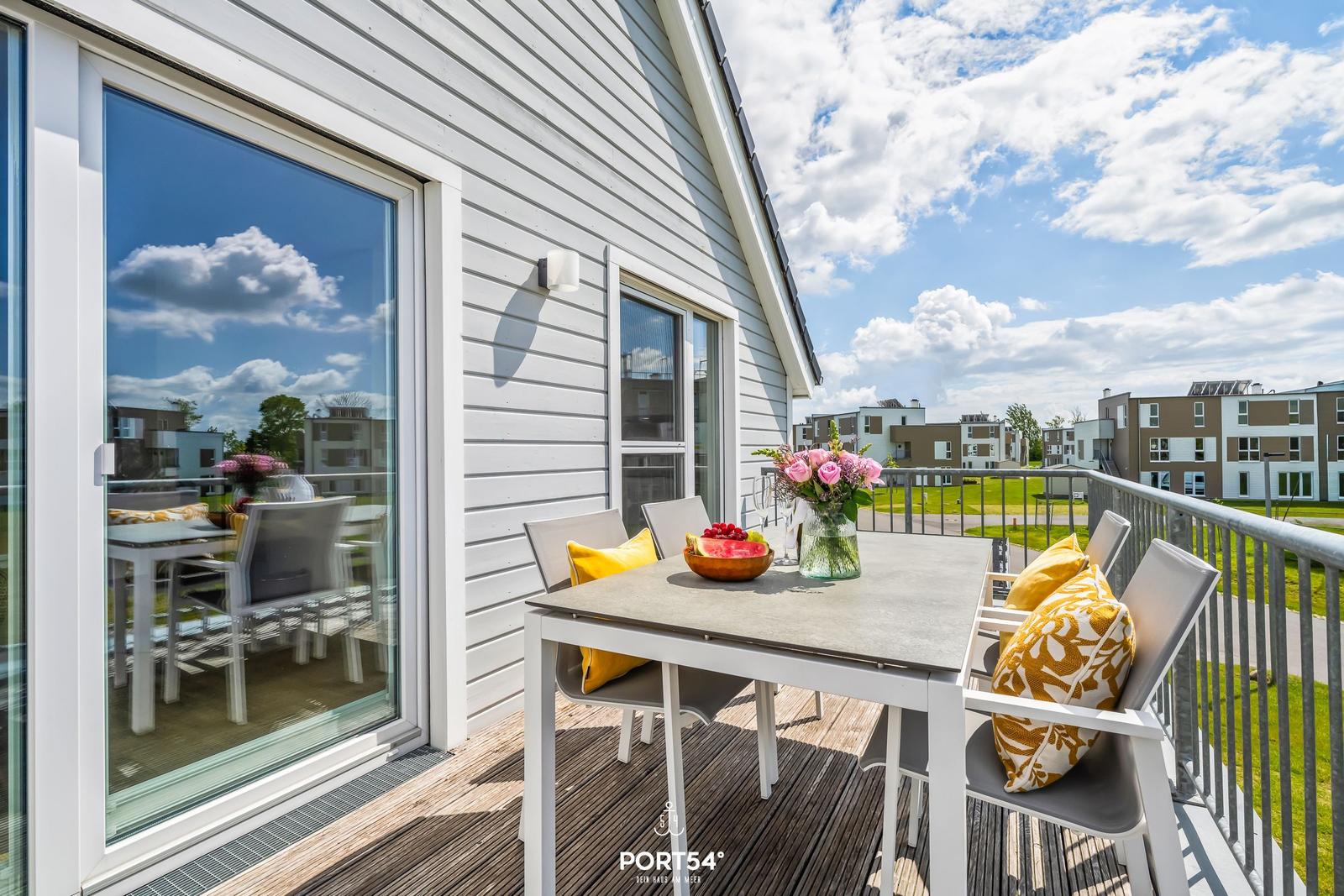 Terrasse mit Tisch, Stühlen und Blumen. Hintergrund: Wohnhäuser und Grünfläche.