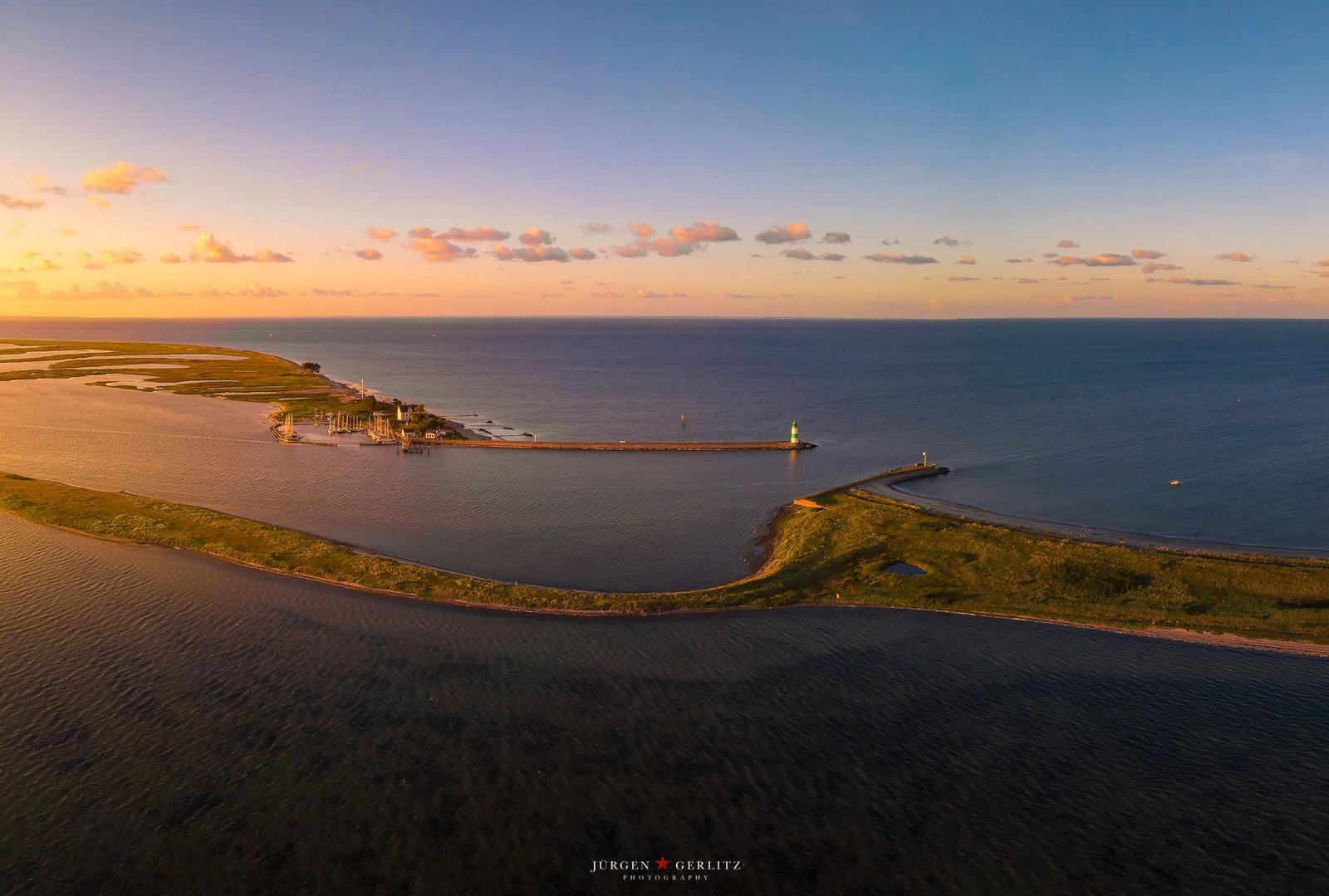 Aerial view of coastal area with lighthouse, marina, and green dunes at sunset.