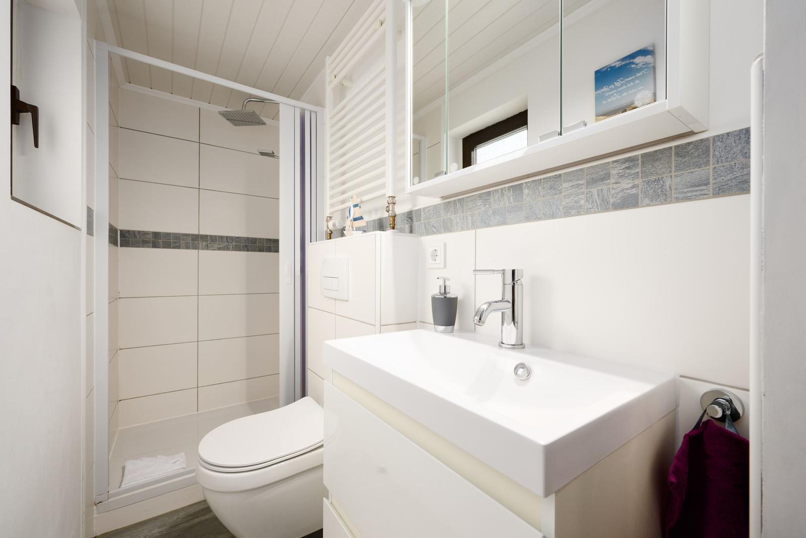 Badezimmer mit Dusche, Waschbecken und Toilette. Weiße Fliesen und Spiegelschrank.