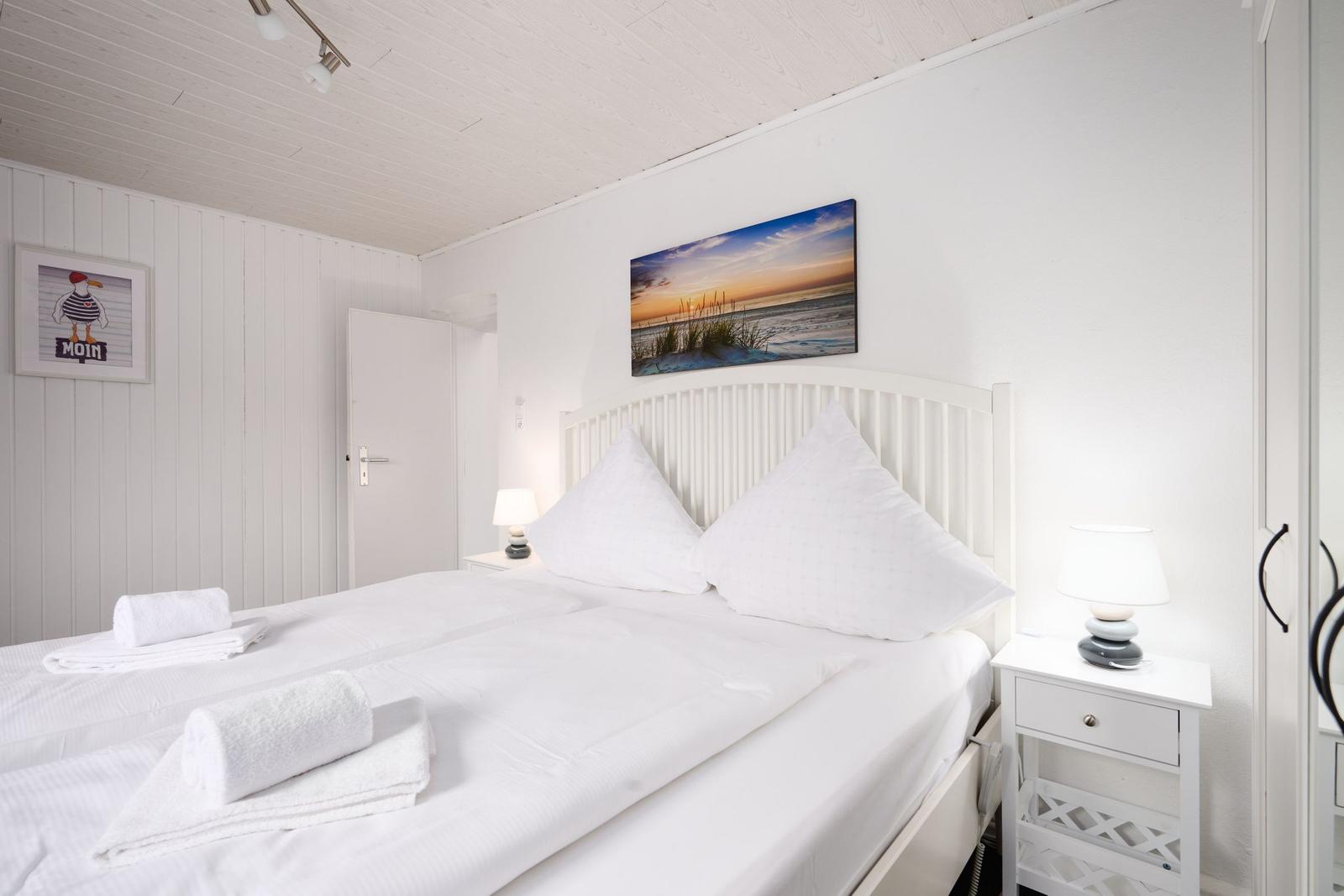 Schlafzimmer mit weißem Bett, Nachttischen und Wandbild. Wand mit vertikalen Paneelen.