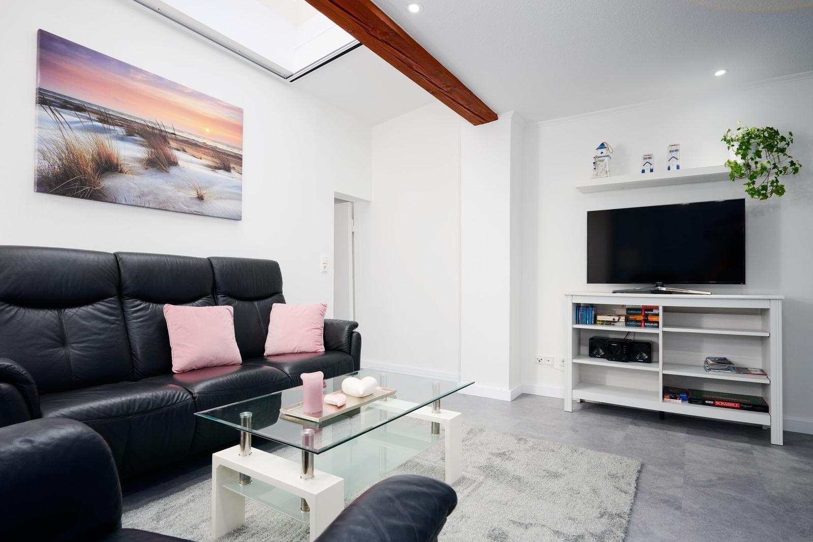 Wohnzimmer mit schwarzer Ledercouch, Glas-Tisch und Fernseher auf weißen Regalen.